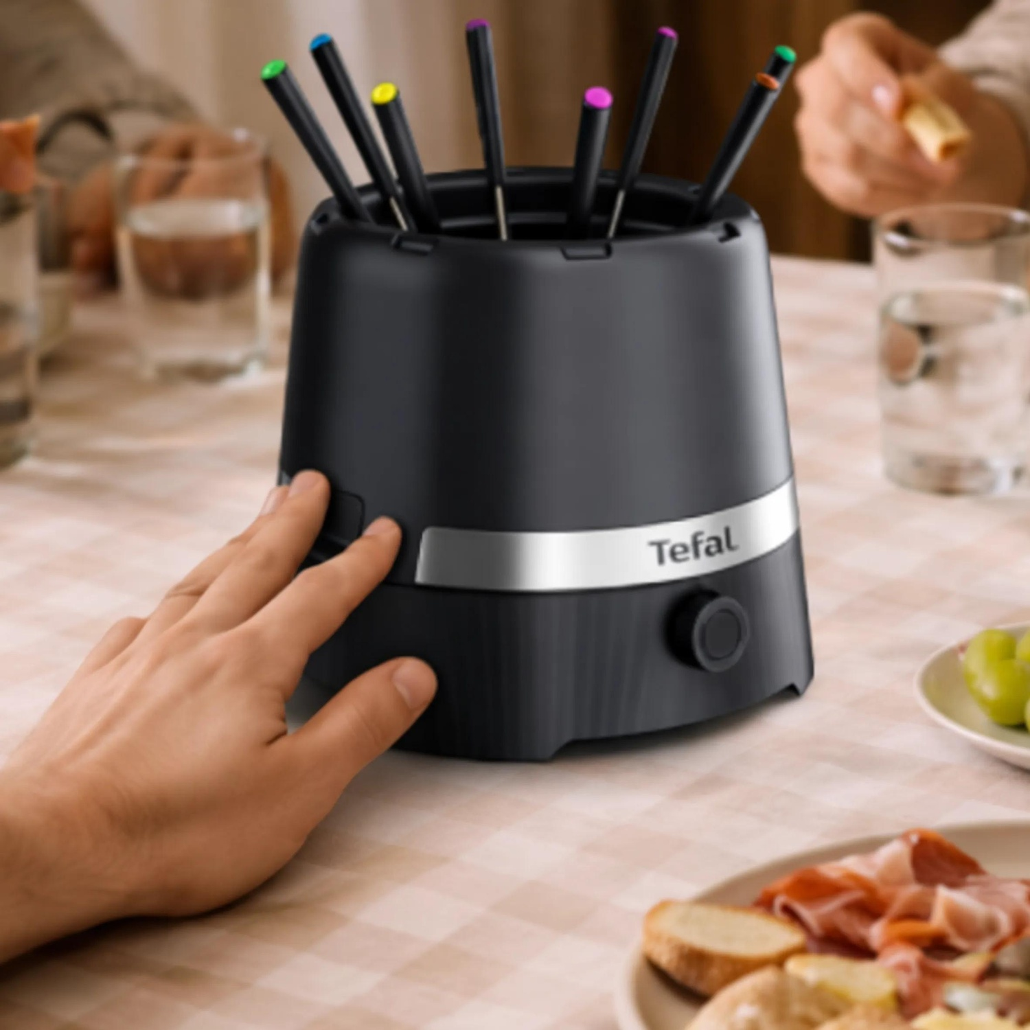 Black Tefal fondue set on a laid table with colorful forks, surrounded by food items and glasses.