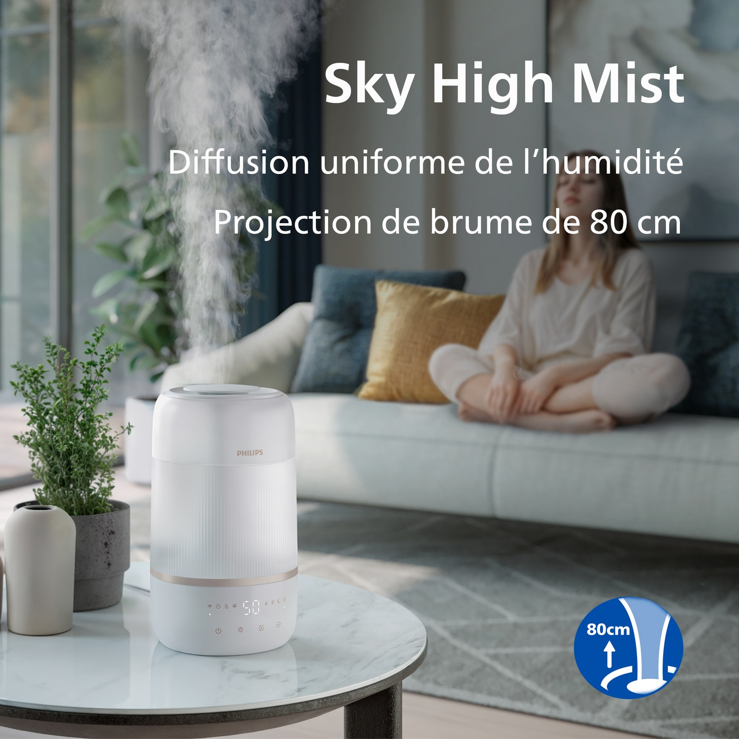 Un humidificateur Philips blanc sur une table dégage de la brume, avec une femme assise sur un canapé.