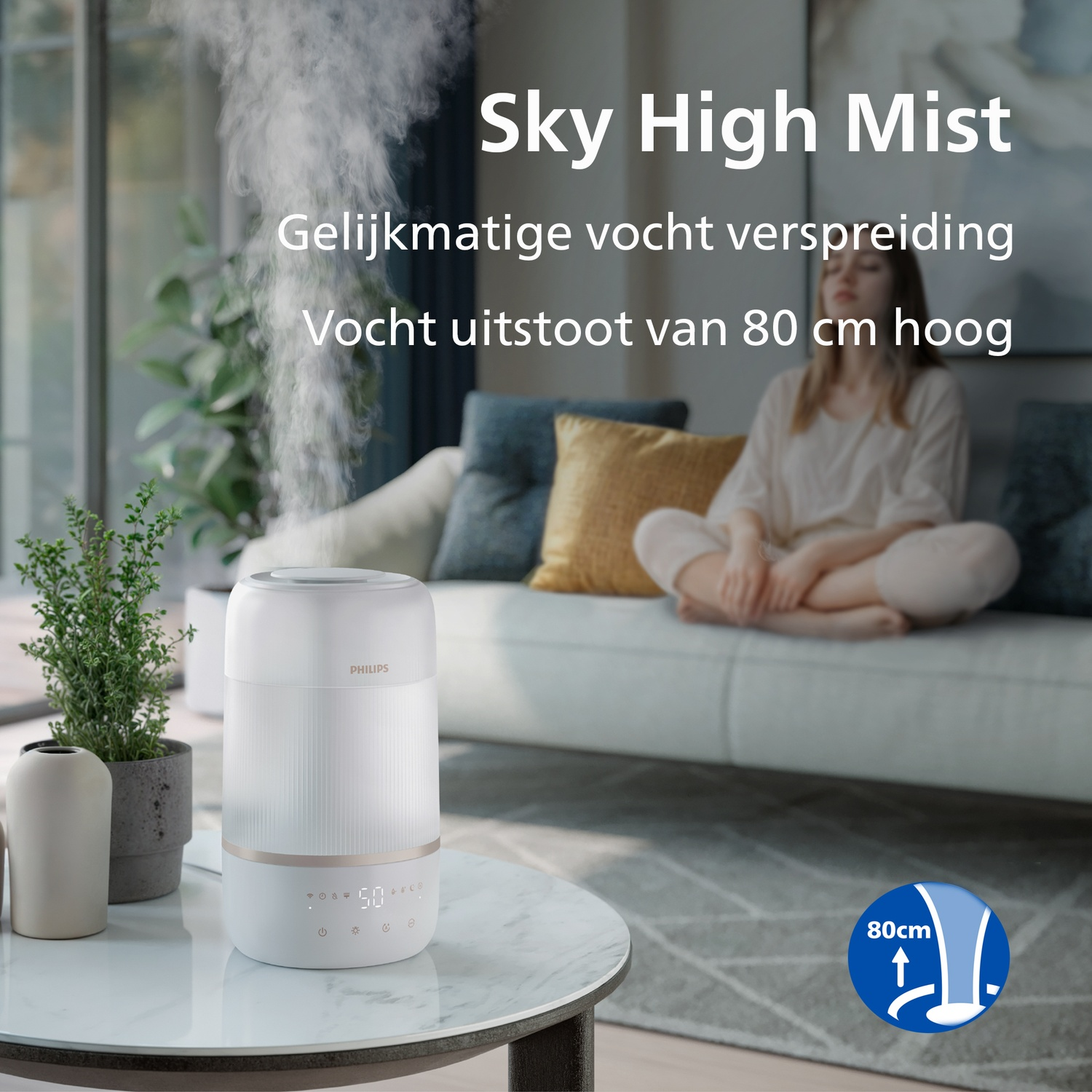 A white Philips humidifier on a table releases tall mist, with a woman meditating on a sofa in the background.