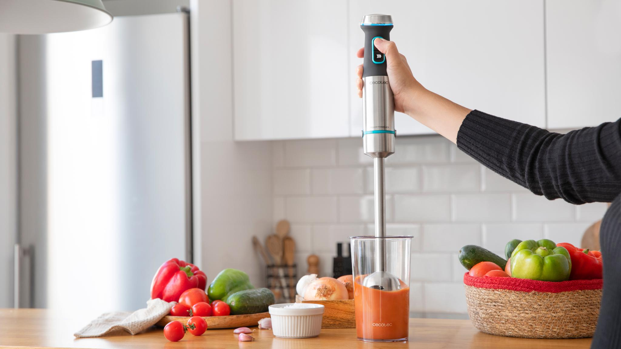 Una mano sostiene una batidora de mano en un vaso con líquido rojo, en una cocina con verduras.