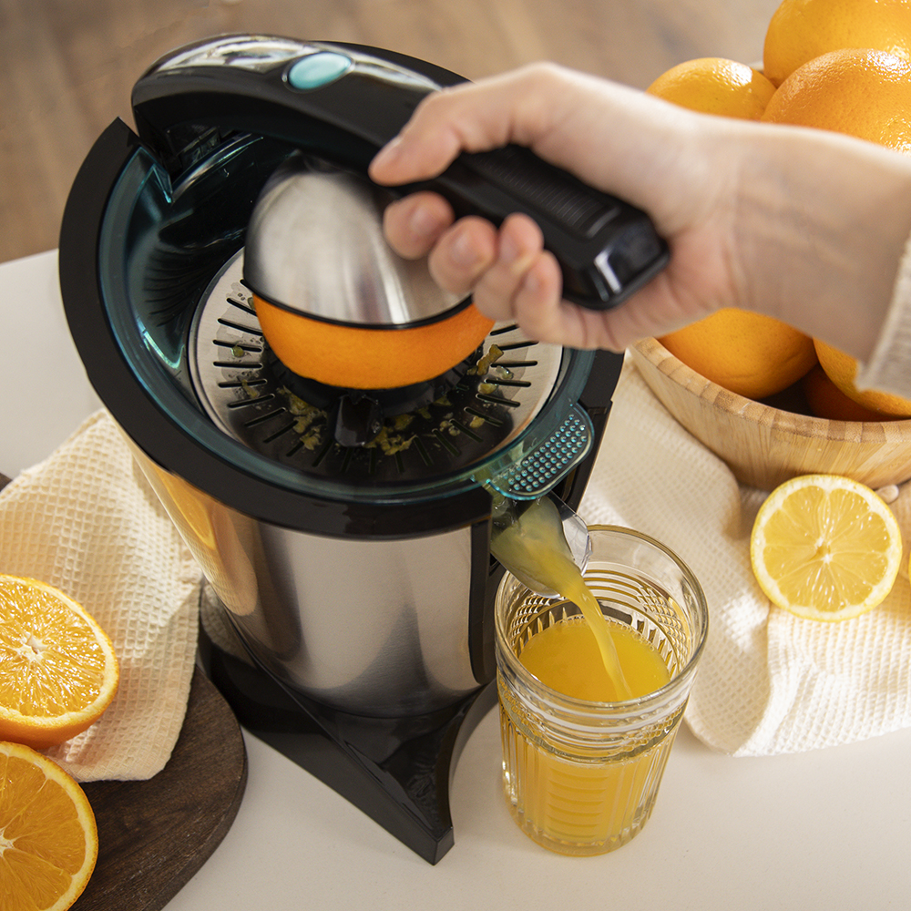 Una mano exprime una naranja en un exprimidor eléctrico, jugo fresco cae en un vaso.