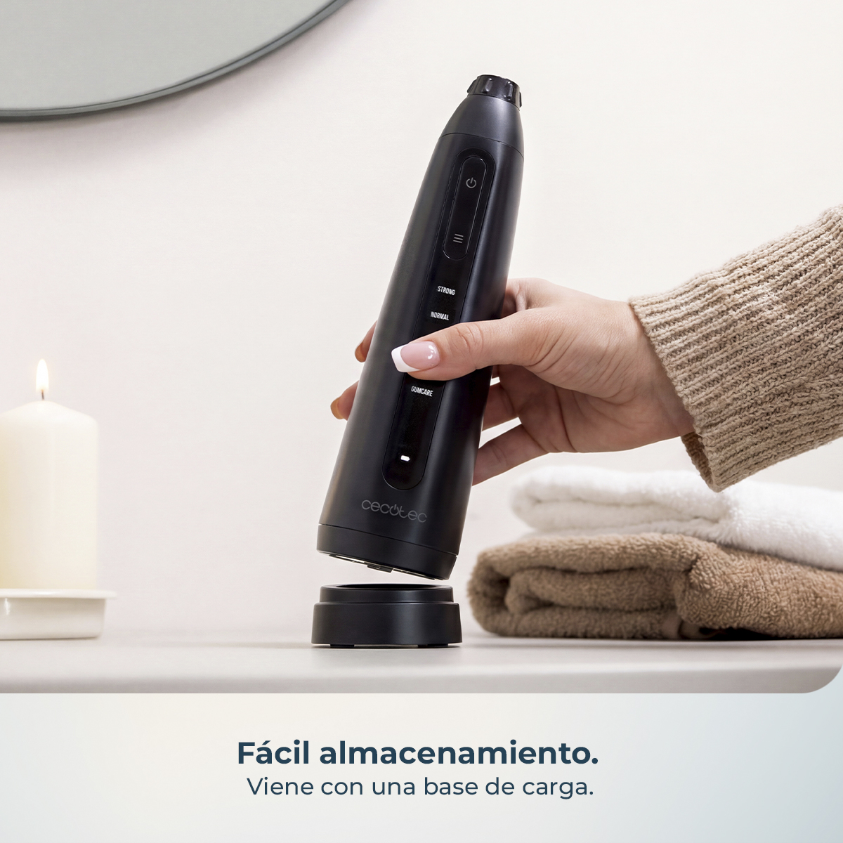Una mano sostiene un limpiador dental eléctrico negro sobre una base de carga en un estante de baño con vela y toallas.