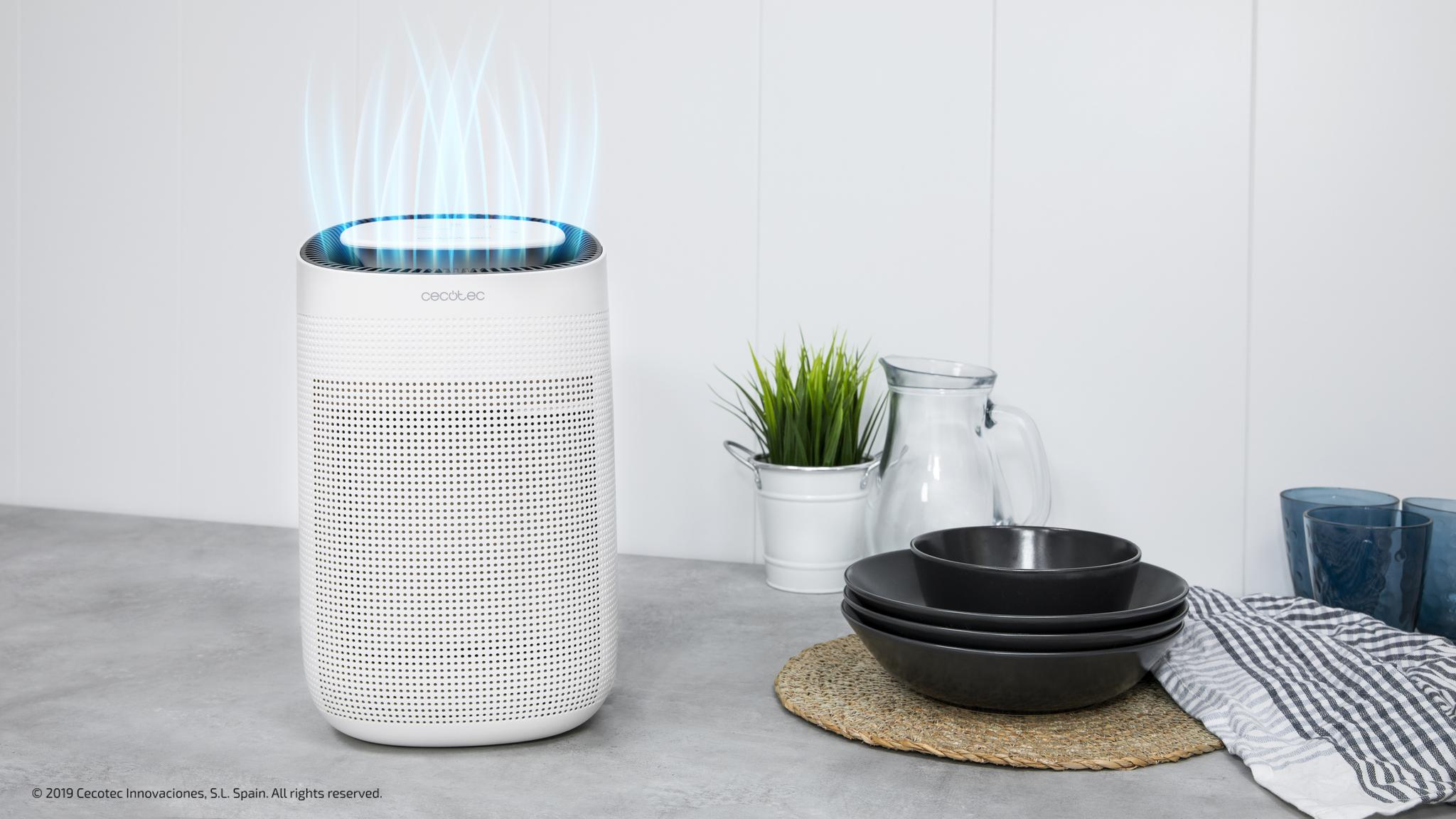 Un purificador de aire blanco en una encimera con luz azul, platos, cuencos y una planta en maceta al fondo.
