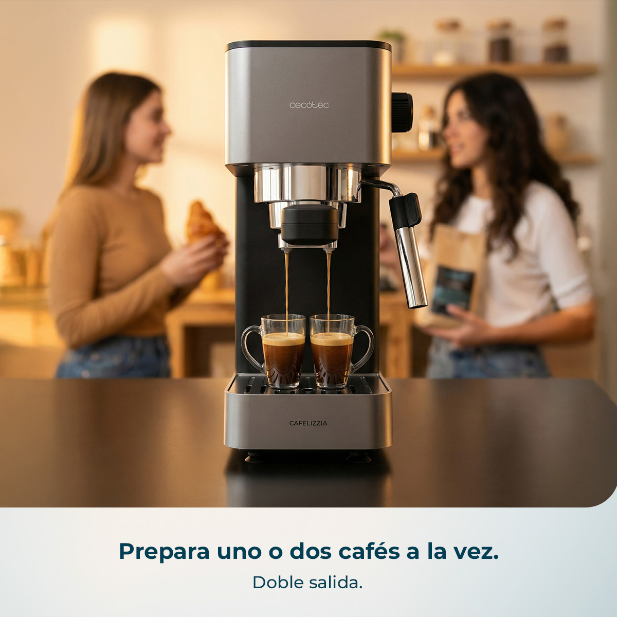 Una cafetera plateada y negra prepara dos tazas de café a la vez, con dos mujeres conversando desenfocadas al fondo.