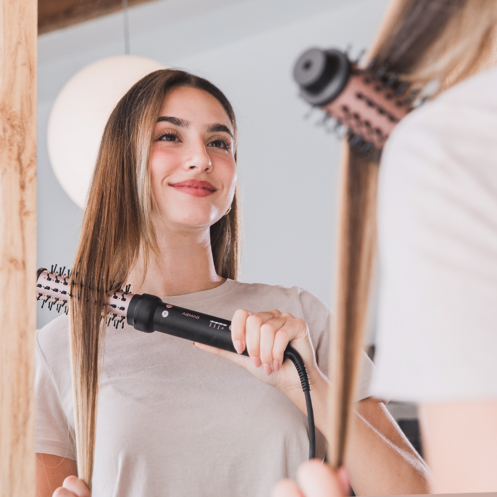 Una mujer sonriente se mira en el espejo y se peina el cabello largo y liso con un cepillo moldeador eléctrico.