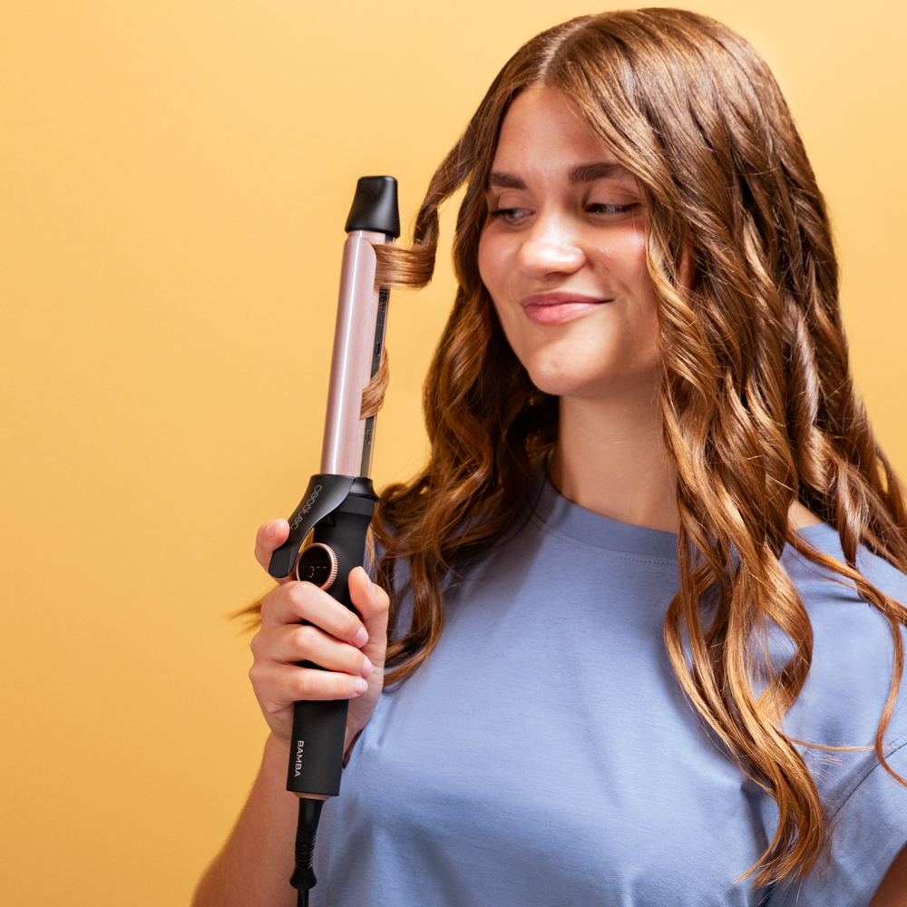 Una mujer sonriente riza su pelo castaño con un rizador rosa y negro sobre fondo amarillo.