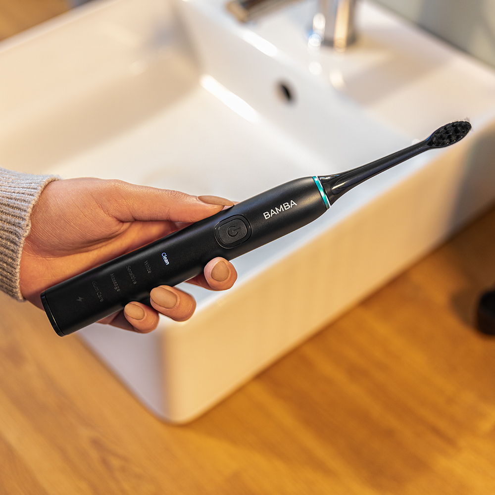 Una mano sostiene un cepillo de dientes eléctrico negro Bamba frente a un lavabo blanco.
