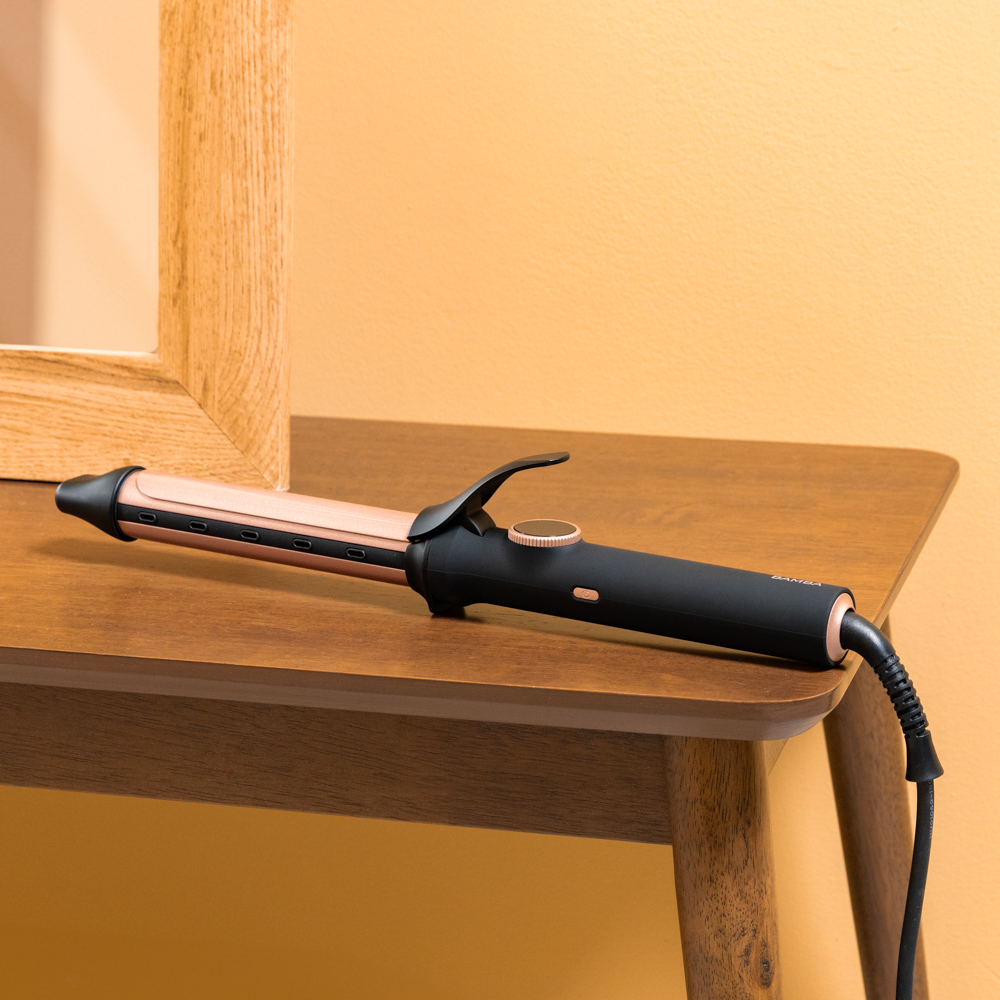 Un rizador de pelo de oro rosa y negro descansa sobre una mesa de madera contra una pared amarilla.