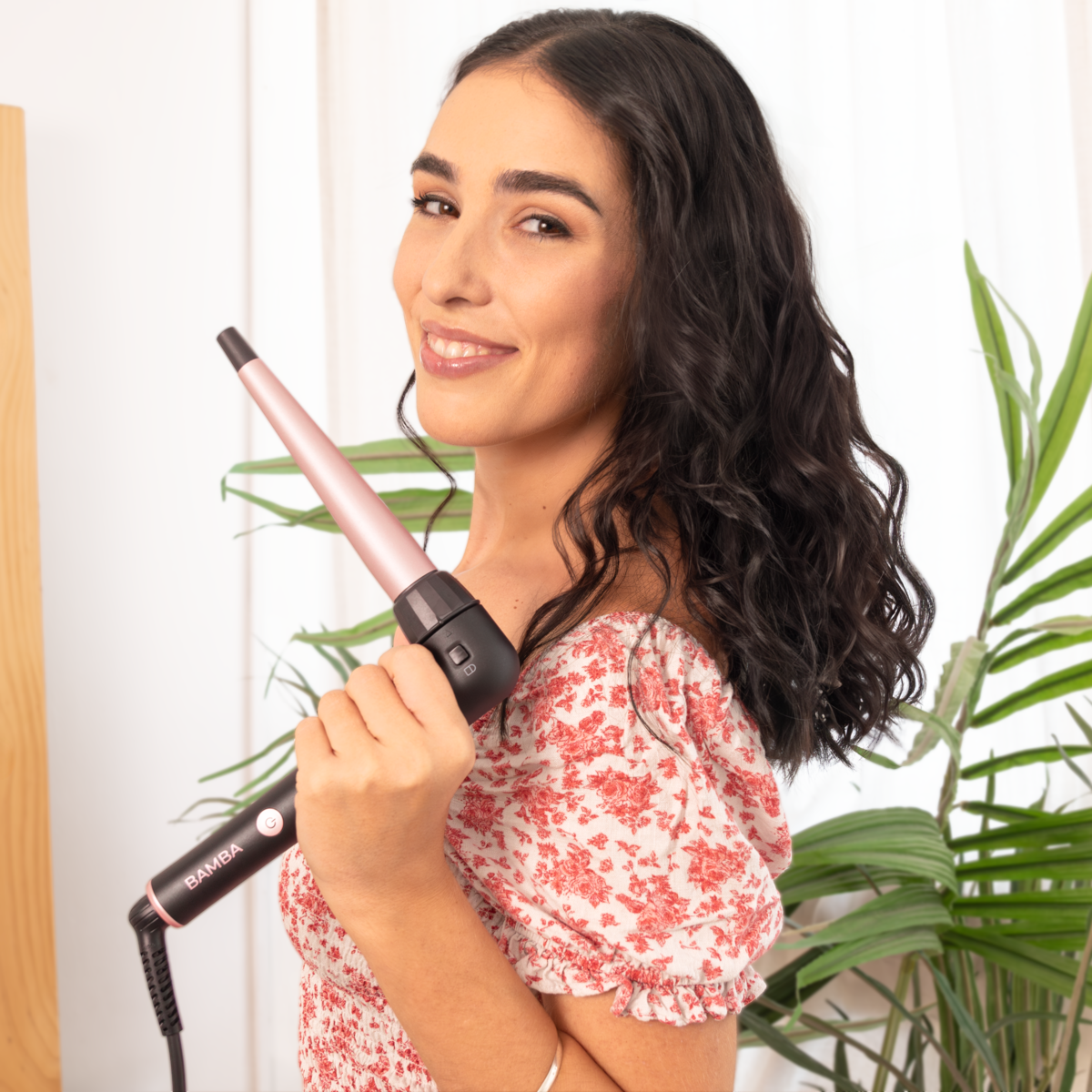 Una mujer sonriente con cabello oscuro sostiene un rizador rosa y negro frente a una planta.