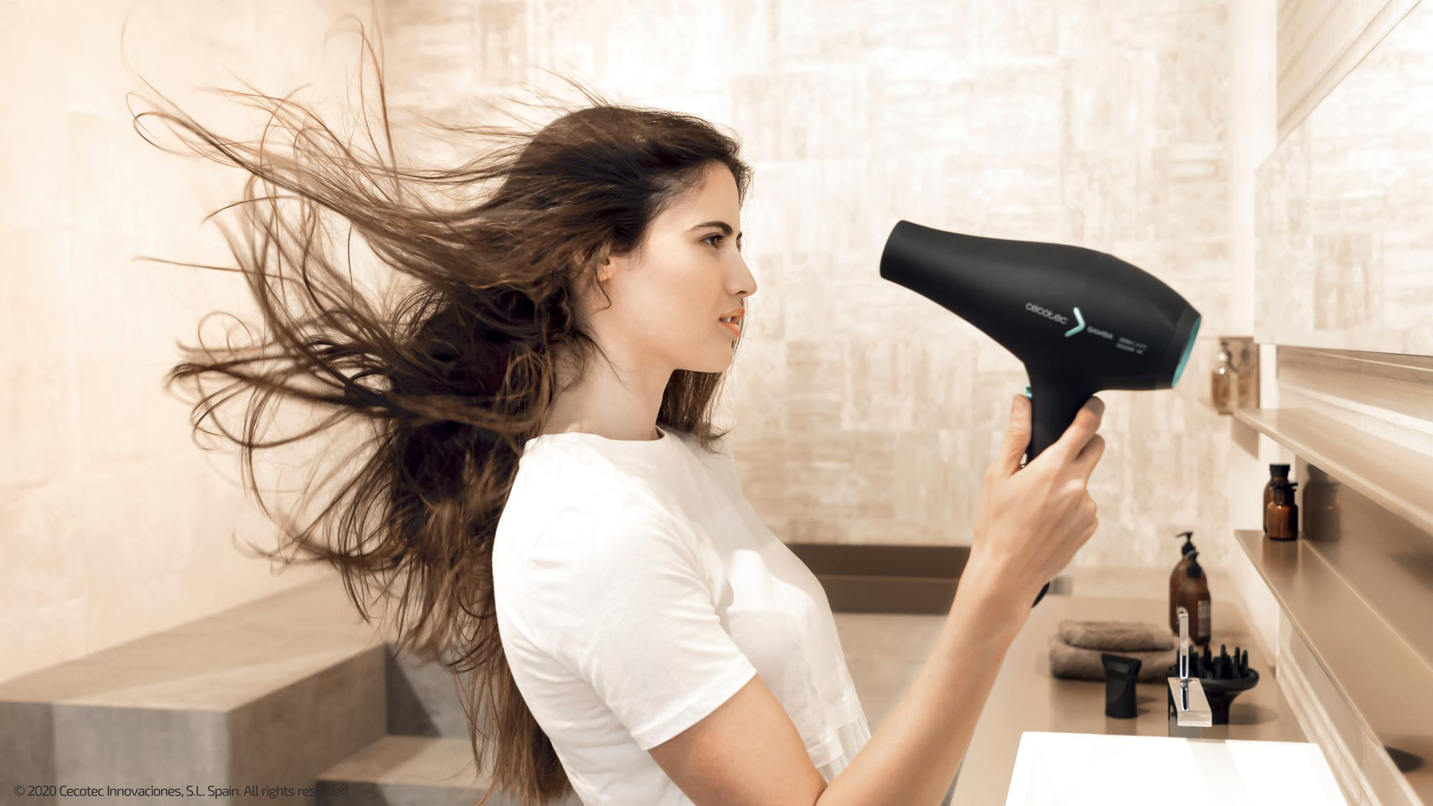 Una mujer con camiseta blanca usa un secador negro en el baño, con su largo cabello castaño al viento.