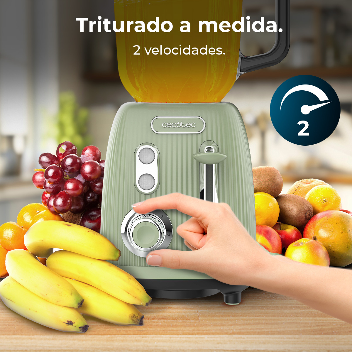 Mano usando licuadora verde frente a fruta con plátanos y uvas.