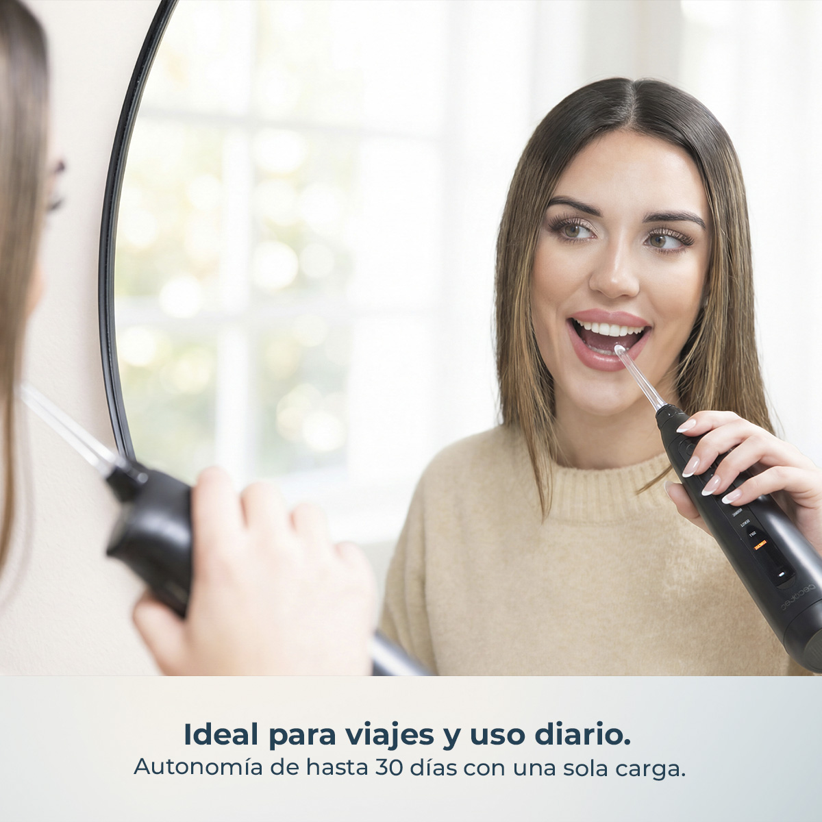 Una mujer sonriente usa un irrigador dental negro frente a un espejo.