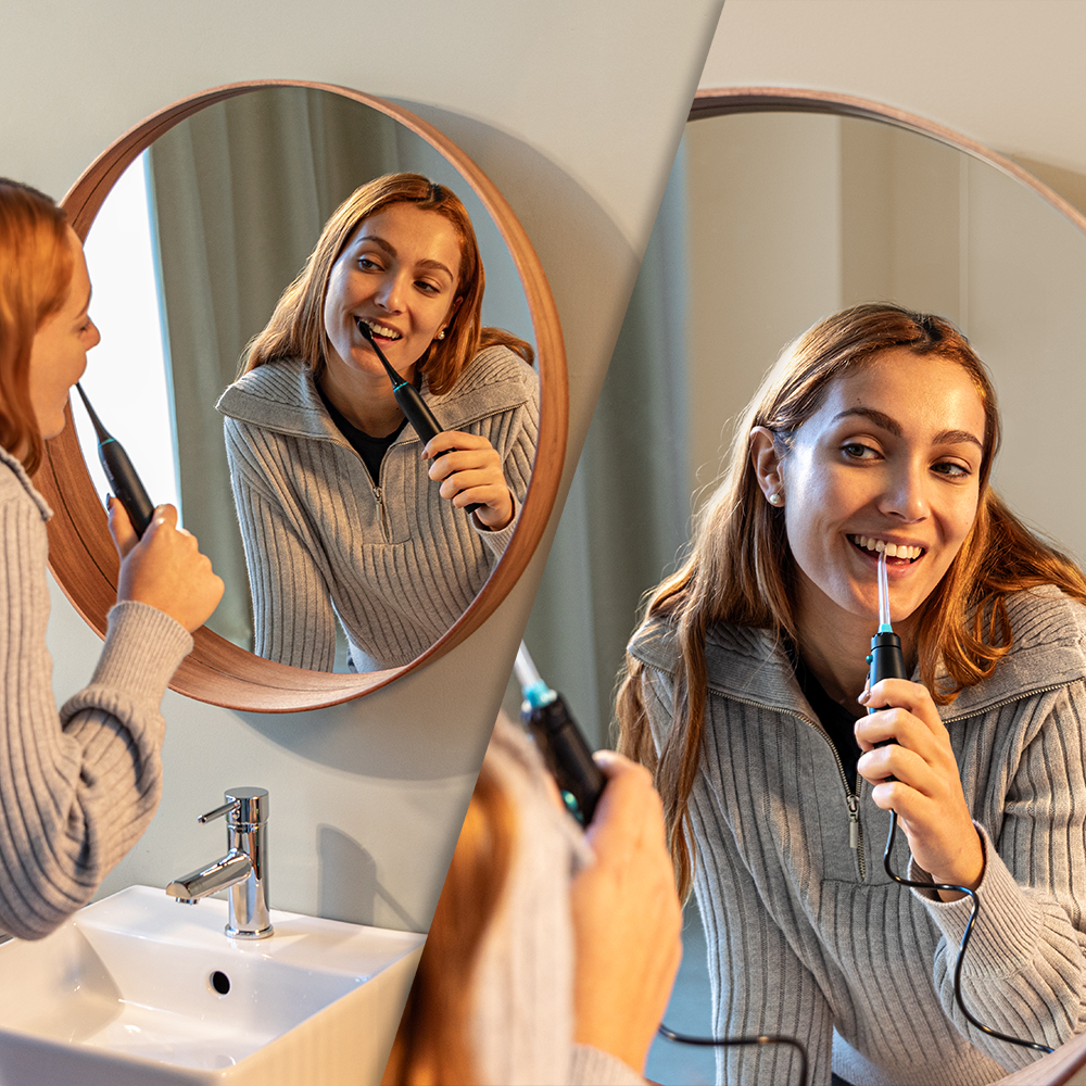 Una mujer con suéter gris se cepilla los dientes con un cepillo eléctrico frente a un espejo de baño redondo.
