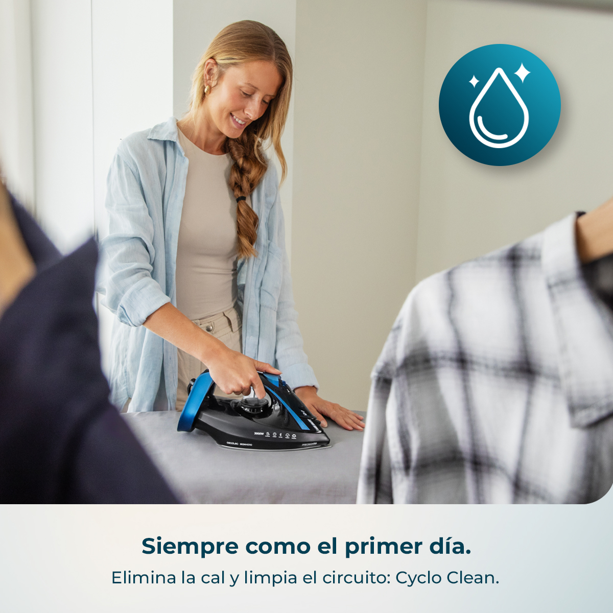 Una mujer sonriente con camisa azul claro plancha ropa con una plancha negra y azul.