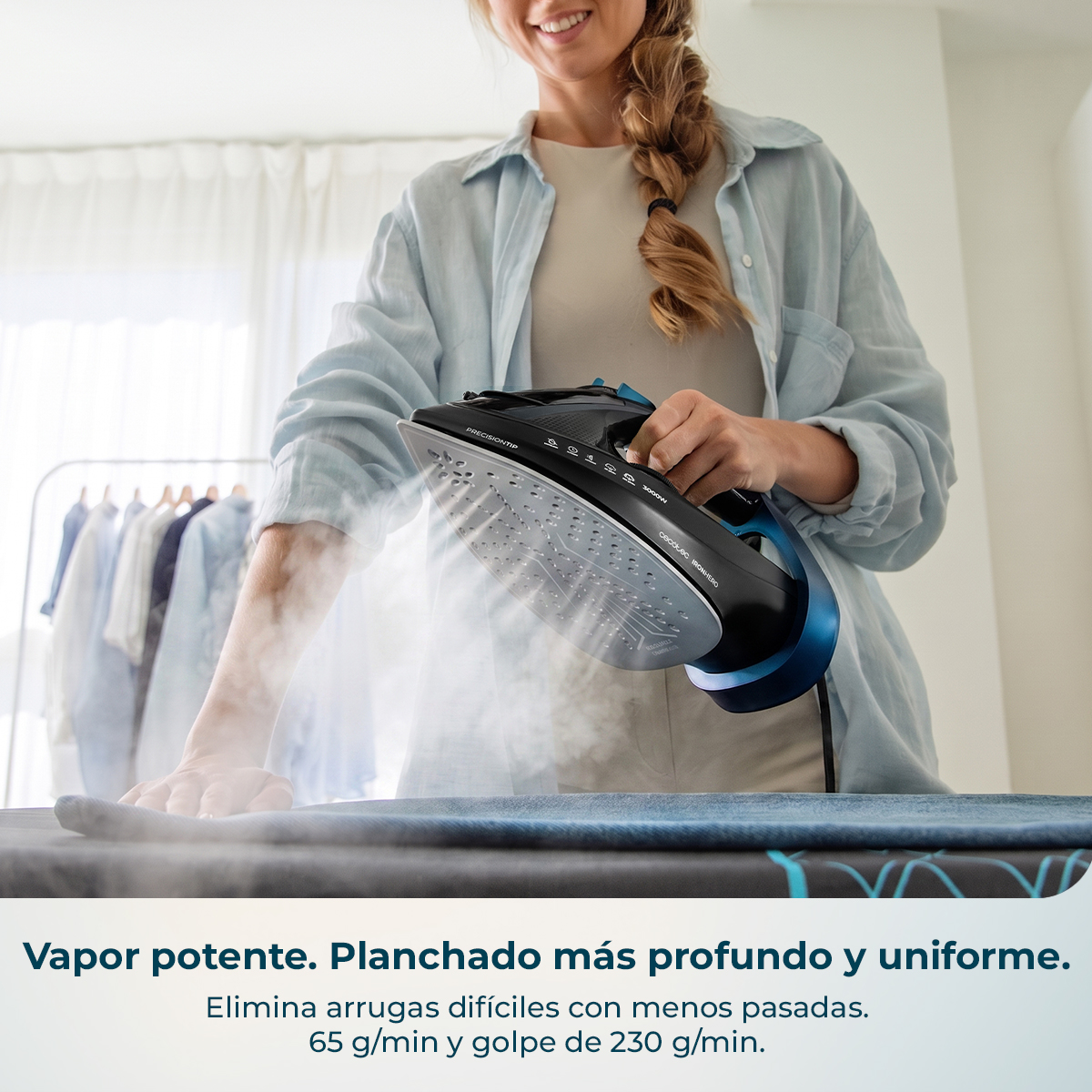 Una mujer planchando ropa en una tabla, sosteniendo una moderna plancha de vapor negra y azul que libera vapor.