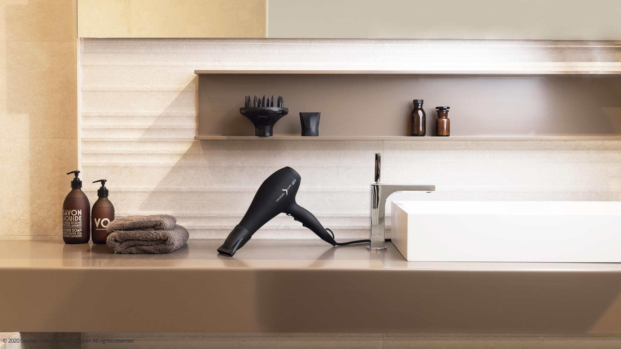 Un secador de pelo negro descansa en un tocador de baño moderno junto a un lavabo blanco, dispensadores de jabón y toallas.