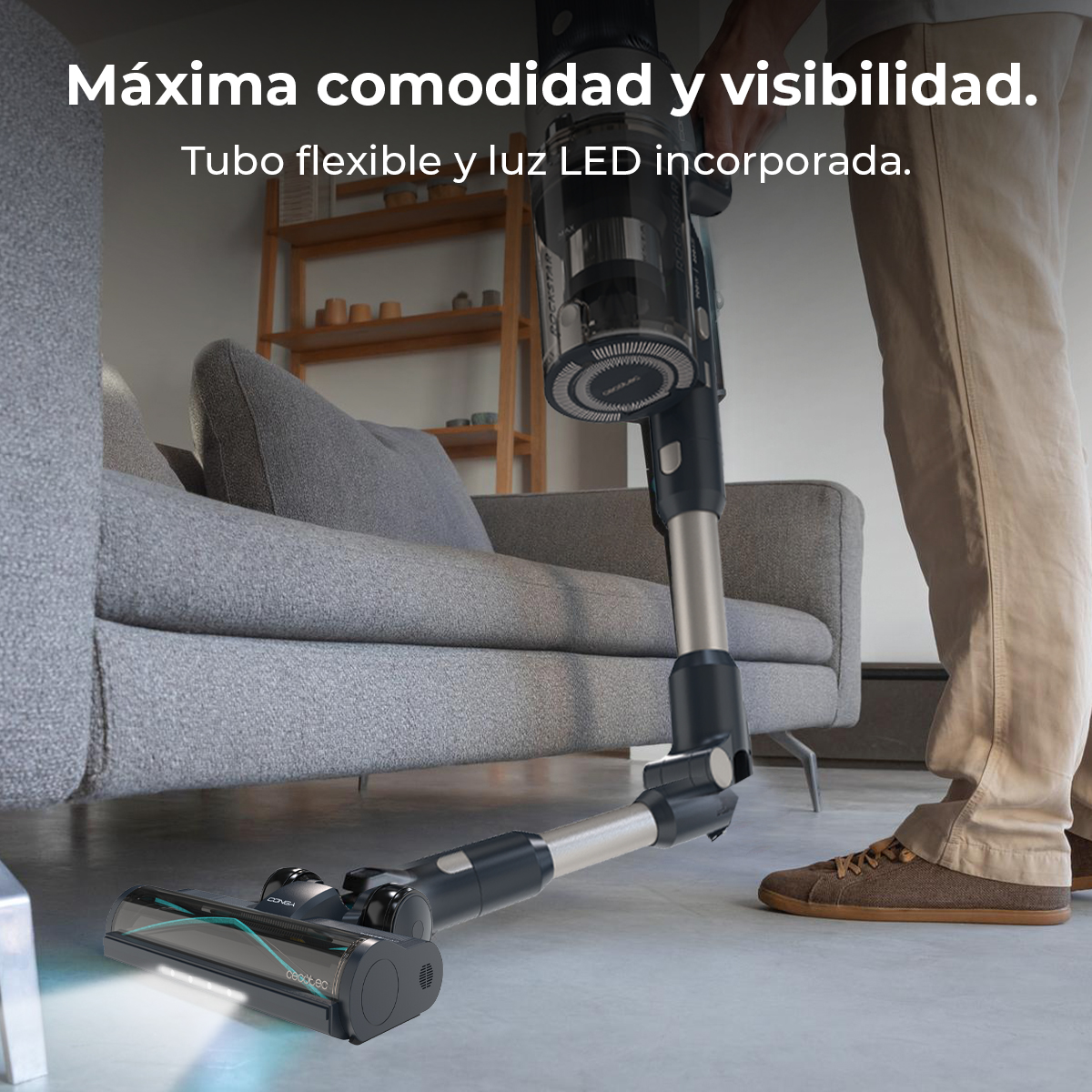 Una persona usa una aspiradora escoba con tubo flexible y luz LED para limpiar bajo el sofá.
