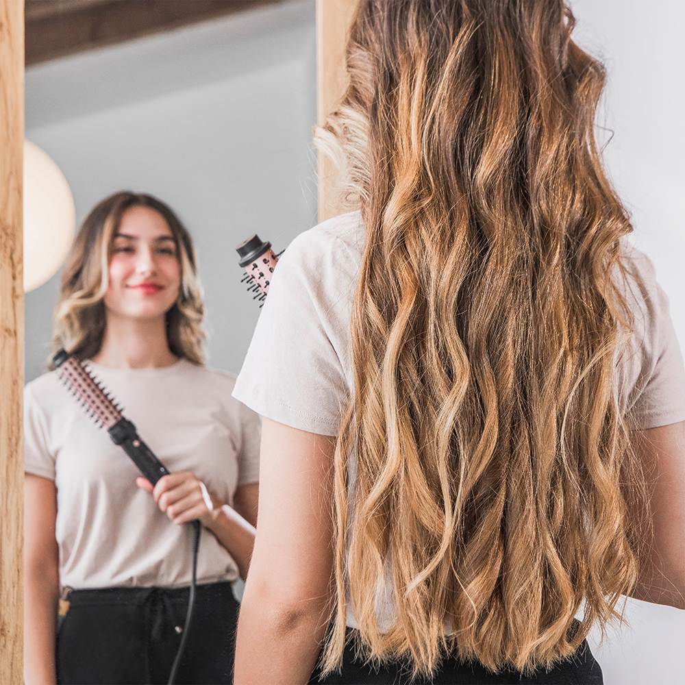 Una mujer con cabello largo y rubio se mira en el espejo sosteniendo un cepillo eléctrico.