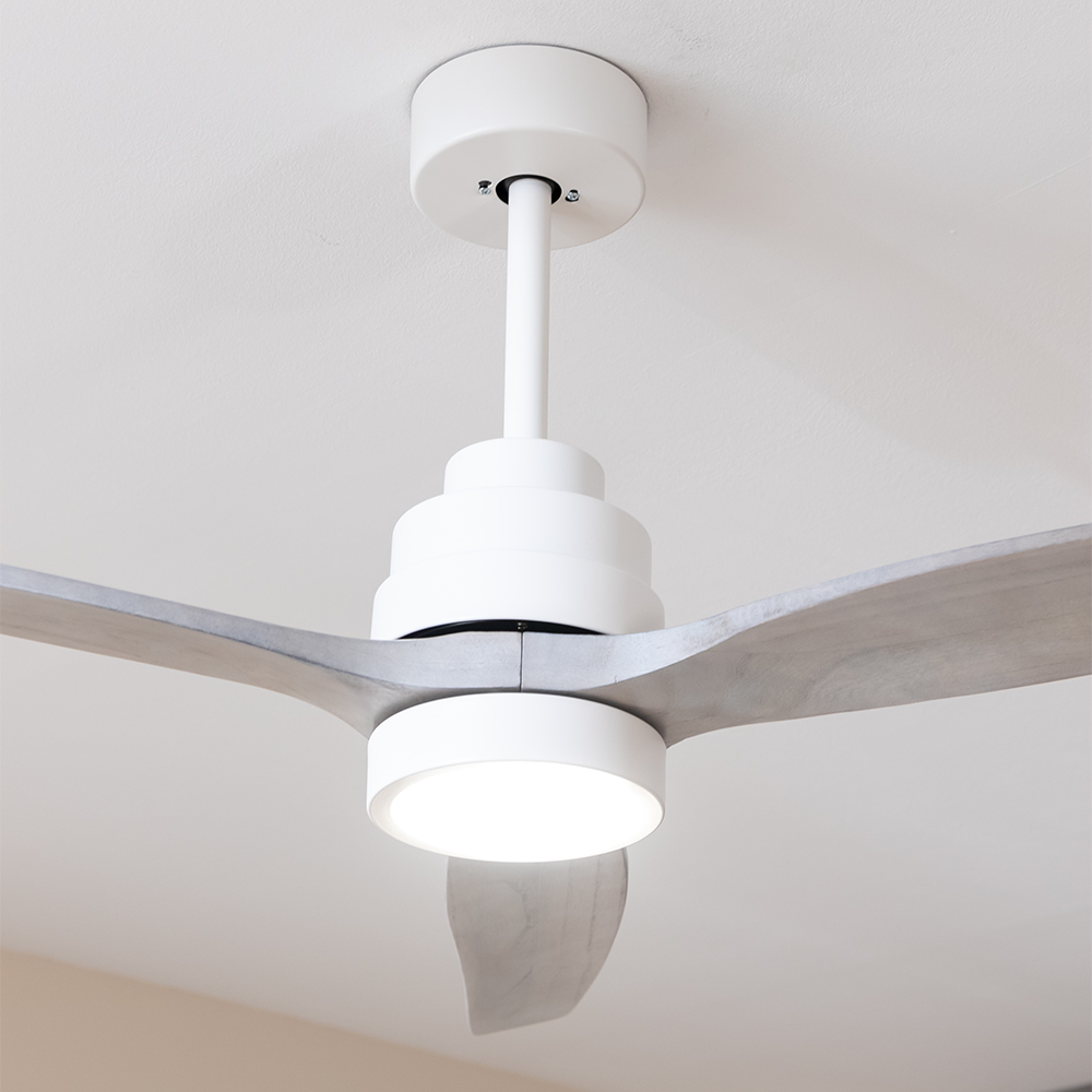 Un moderno ventilador de techo blanco con aspas de madera clara y luz central brillante en un techo blanco.
