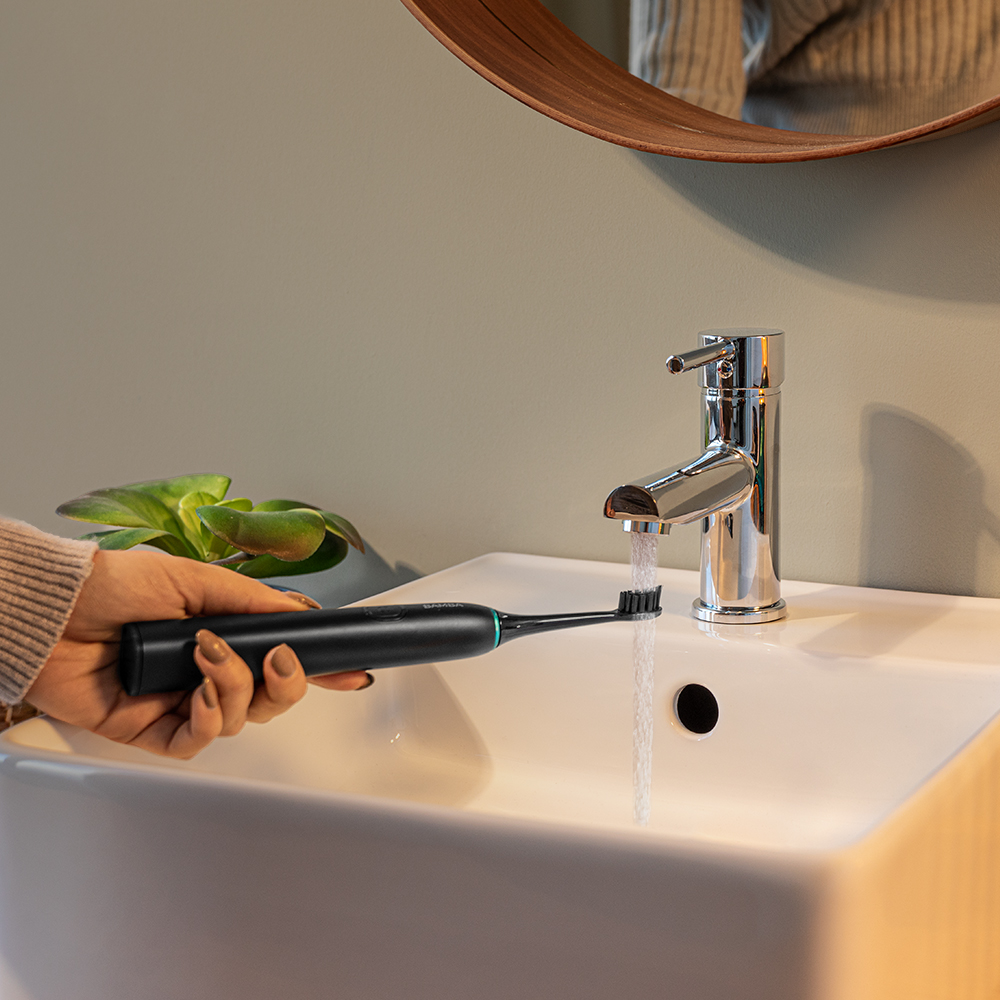 Mano sosteniendo un cepillo de dientes eléctrico negro bajo un grifo abierto en un lavabo blanco.