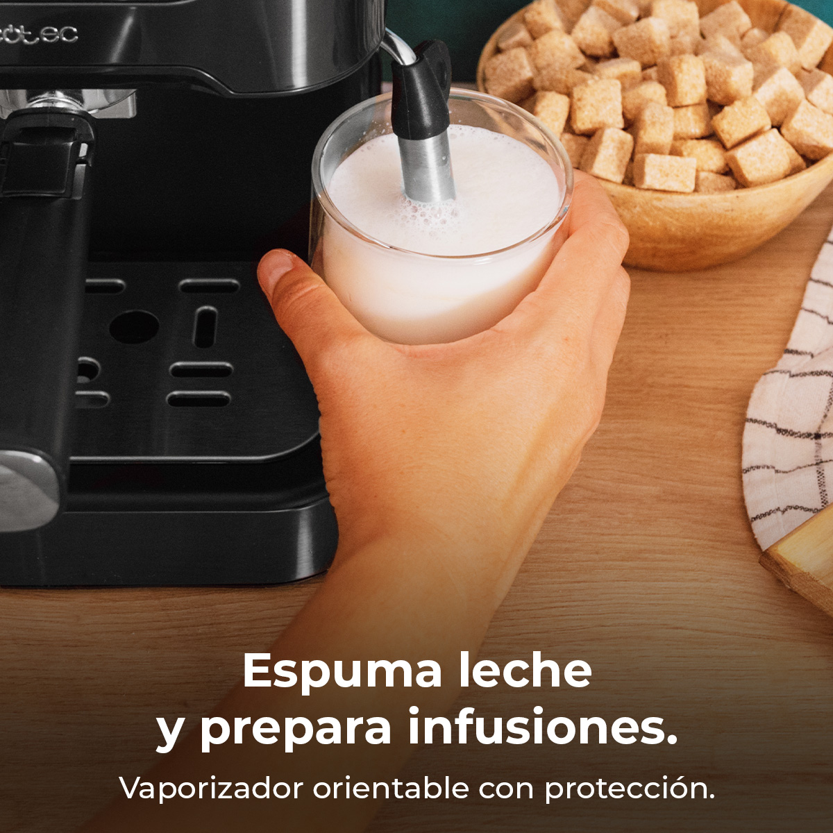 Una mano sostiene un vaso de leche mientras la cafetera la espumea. Un bol de madera con terrones de azúcar al lado.
