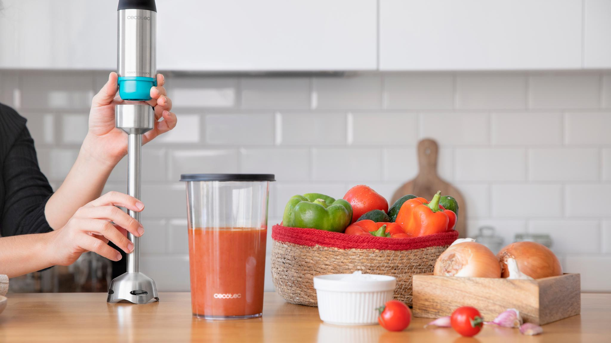 Persona sosteniendo una batidora de mano junto a un vaso de jugo de vegetales y verduras frescas.