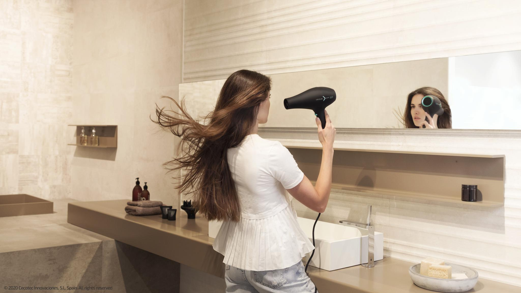 Una mujer con camisa blanca se seca el pelo largo frente a un espejo en un baño moderno.