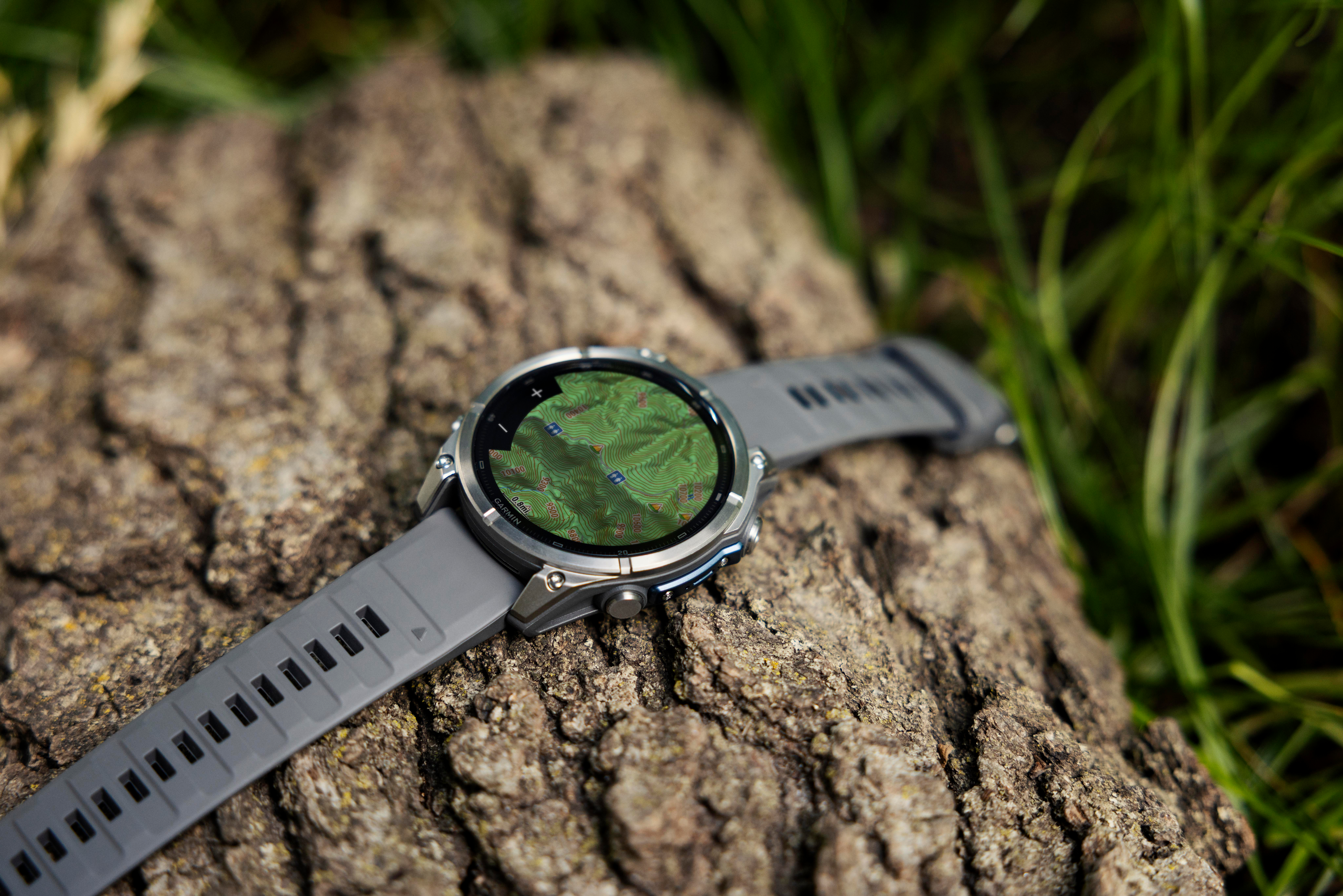 Eine graue Smartwatch mit einer topo-Karte auf dem Display, liegend auf rauer Baumrinde neben grünem Gras.