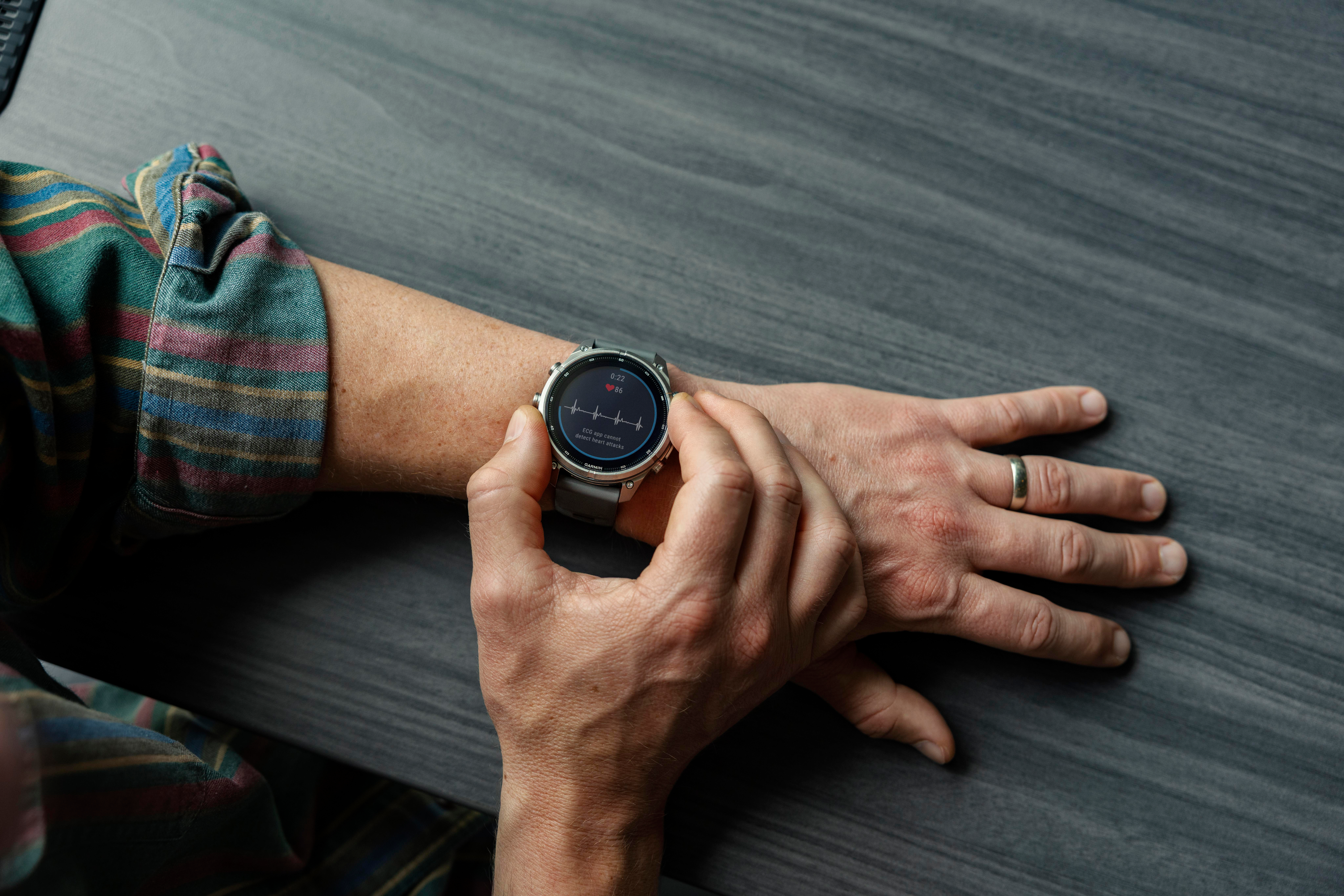 Eine Person bedient mit den Fingern eine Smartwatch mit EKG-Grafik auf einem dunklen Holztisch.