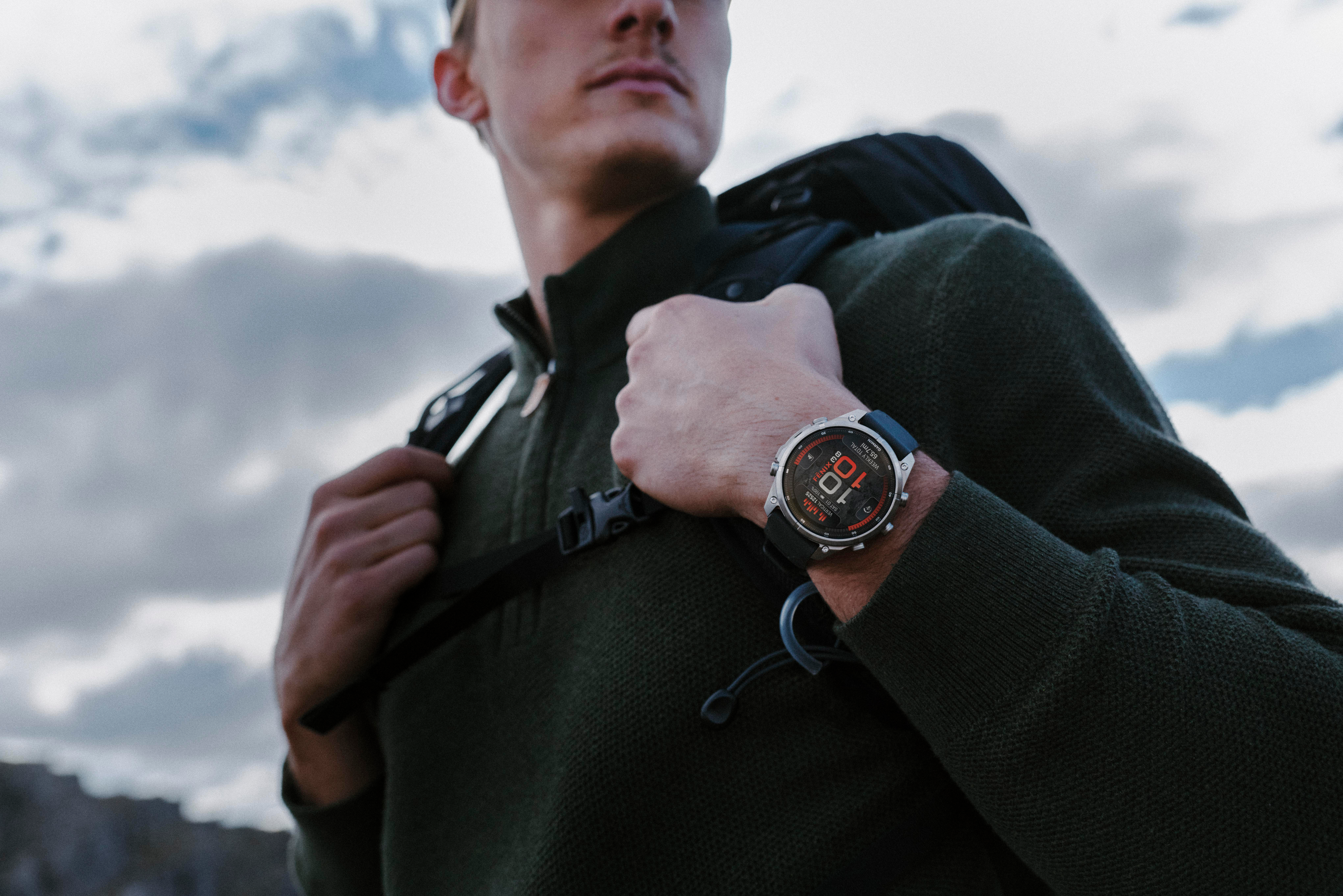 Ein Mann im grünen Pullover mit Rucksack und Outdoor-Uhr am Handgelenk vor bewölktem Himmel.