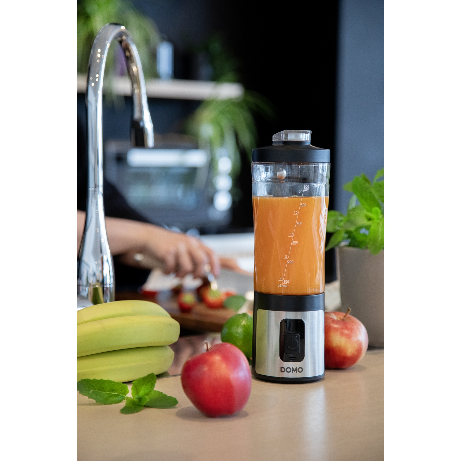 Un blender Domo avec du jus d'orange sur un plan de travail, entouré de pommes, de bananes et d'un robinet.