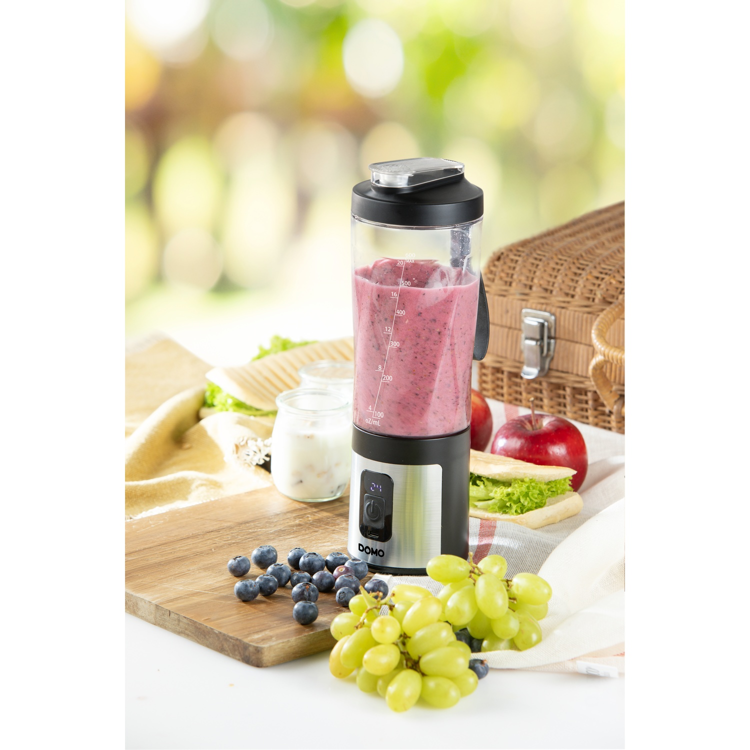 Un blender avec un smoothie rose, entouré de raisins, myrtilles, sandwichs et un panier de pique-nique sur une planche.