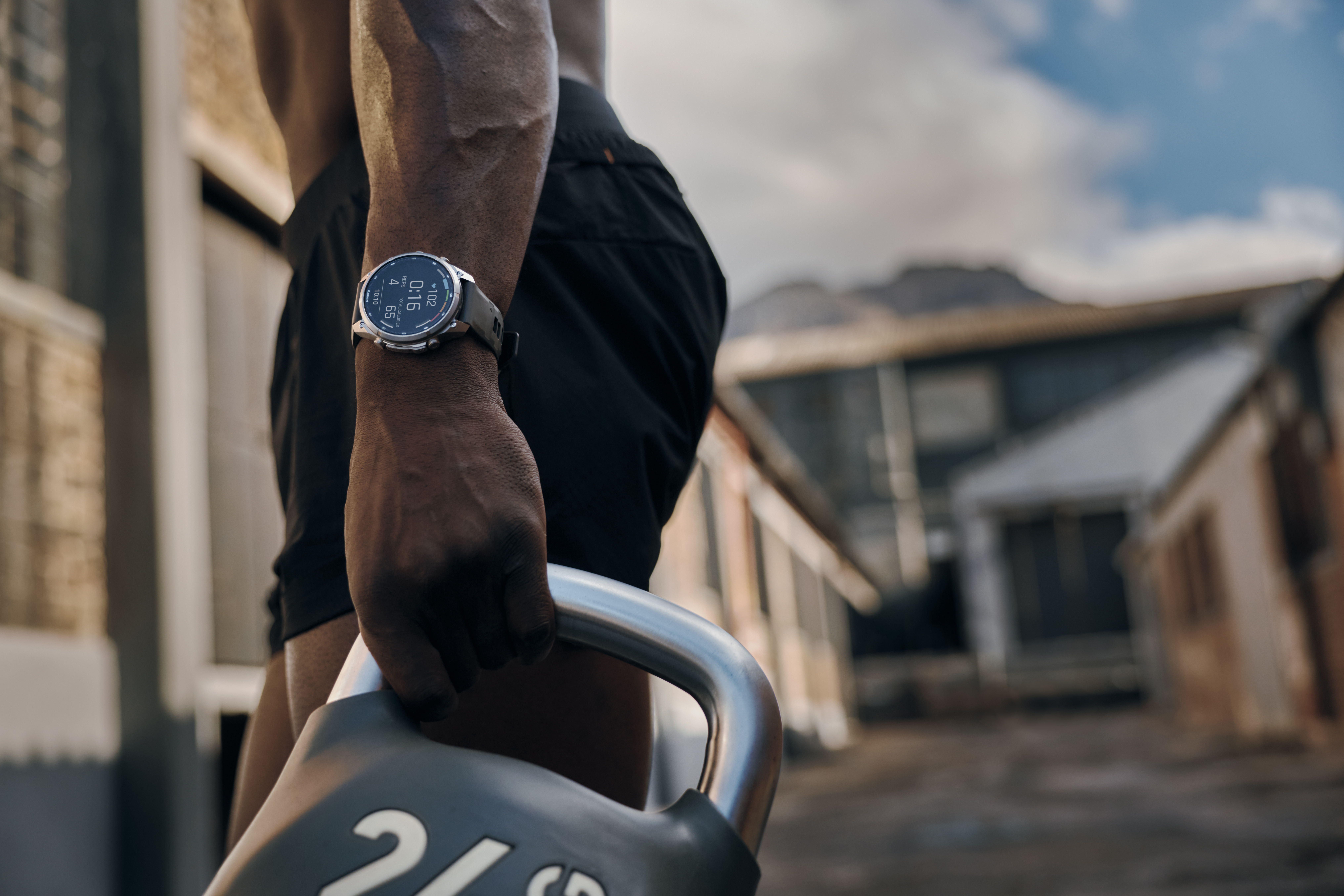 Ein Sportler mit Fitness-Uhr hält eine schwere Kettlebell in der Hand vor einem verschwommenen Gebäude.