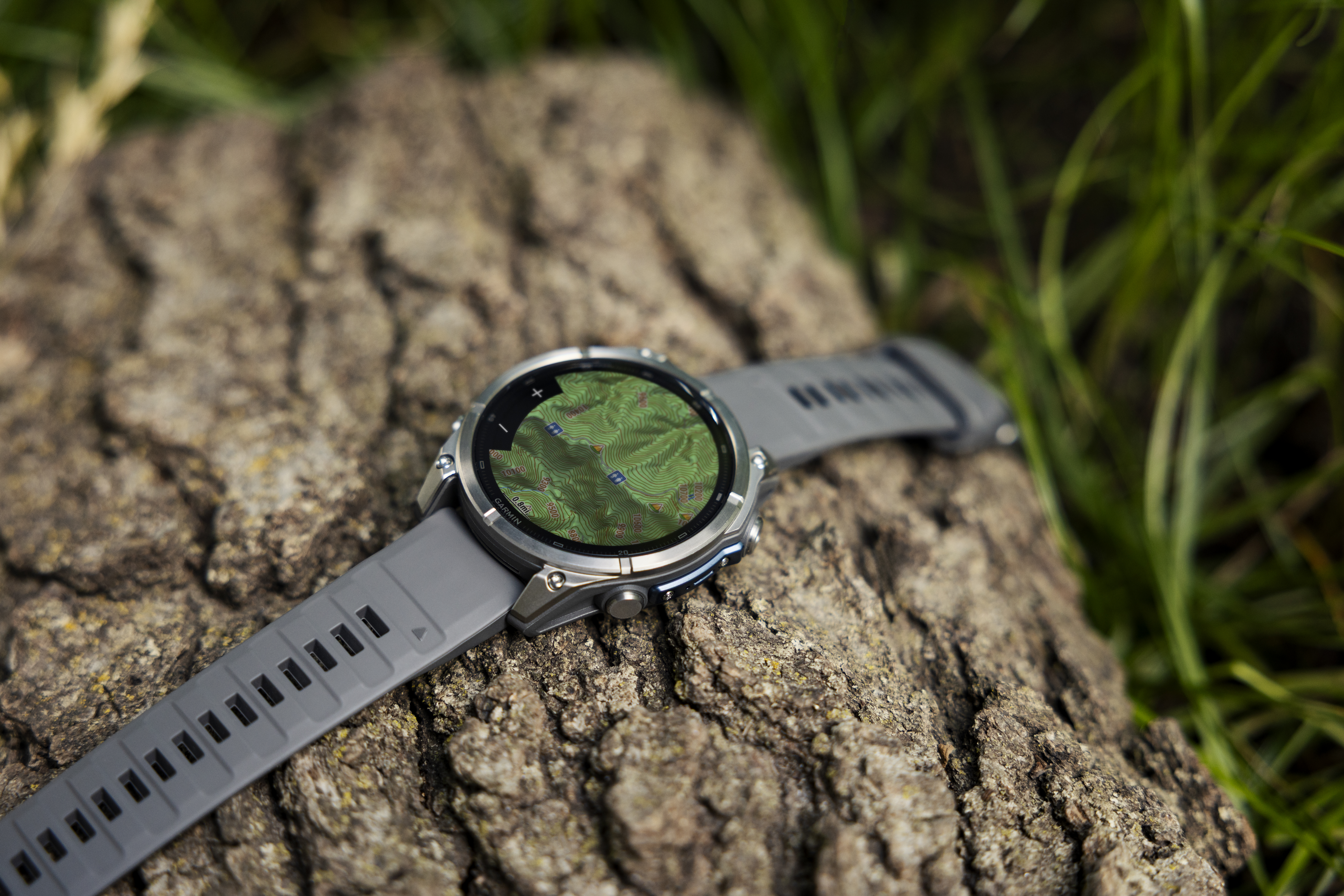 Eine graue Smartwatch mit topografischer Karte auf dem Display liegt auf rauer Baumrinde, umgeben von grünem Gras.