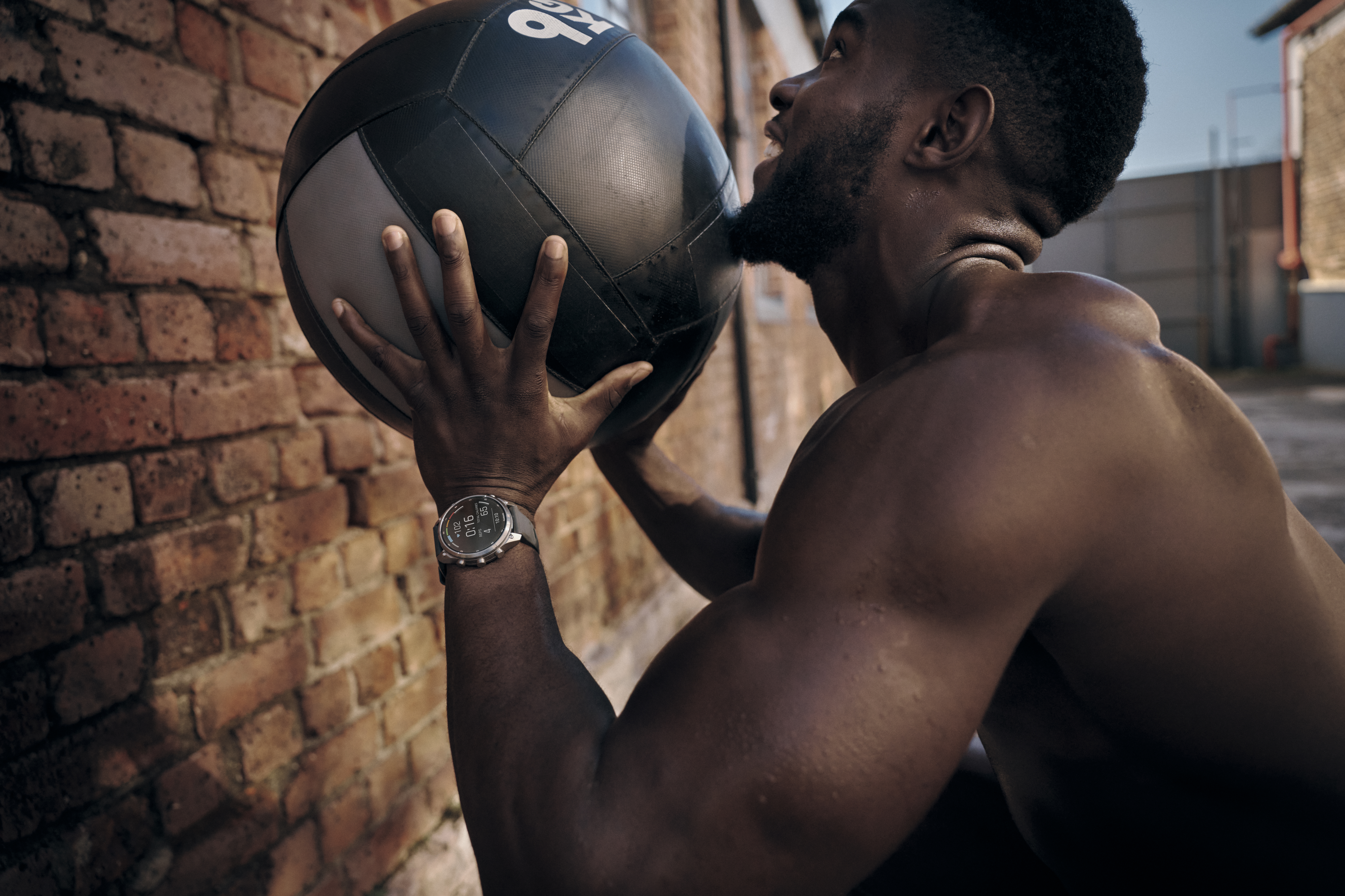 Ein athletischer Mann mit Sportuhr hält vor einer Ziegelwand einen Medizinball.