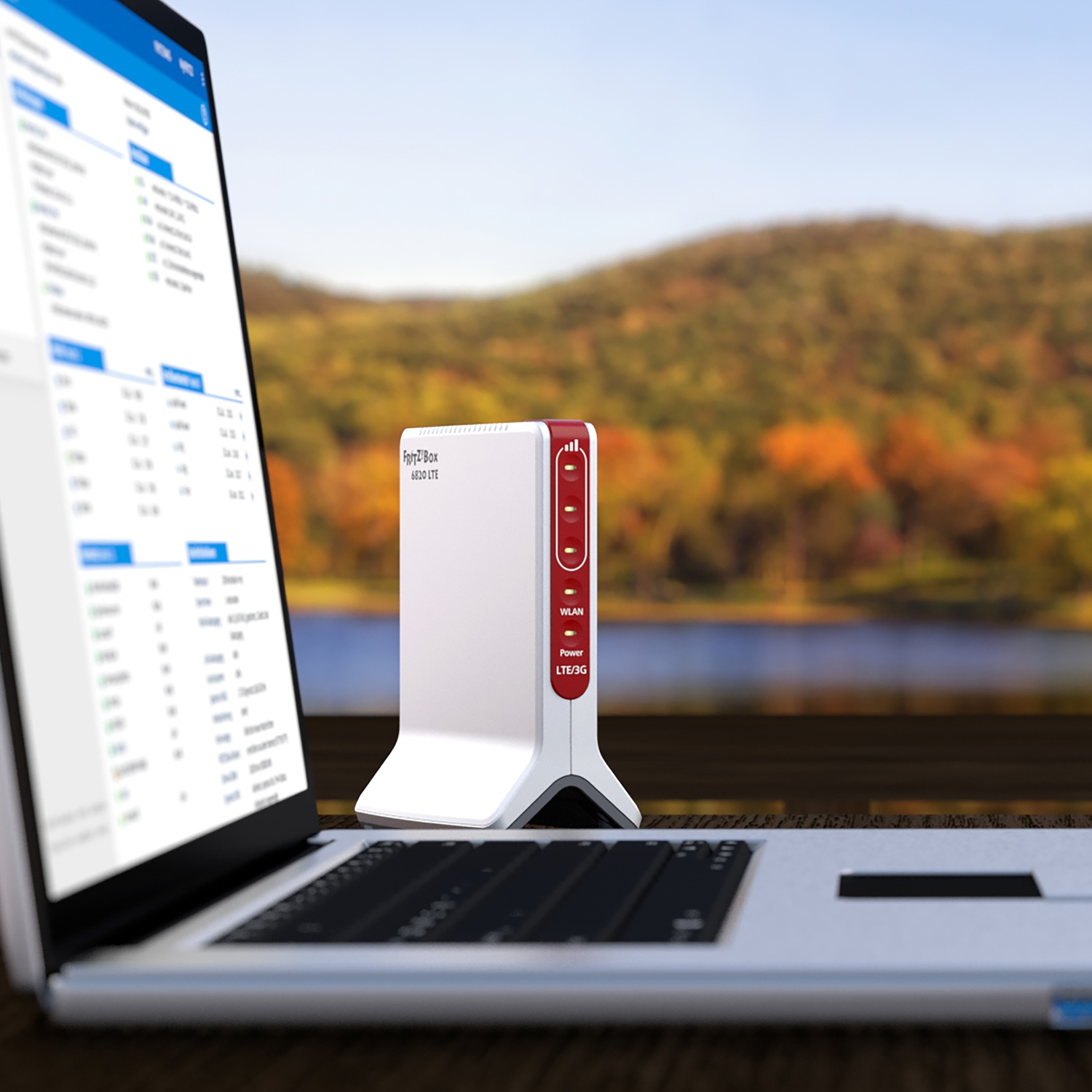 Un router LTE bianco e rosso accanto a un computer portatile su un tavolo con sfondo di montagne e lago autunnale.