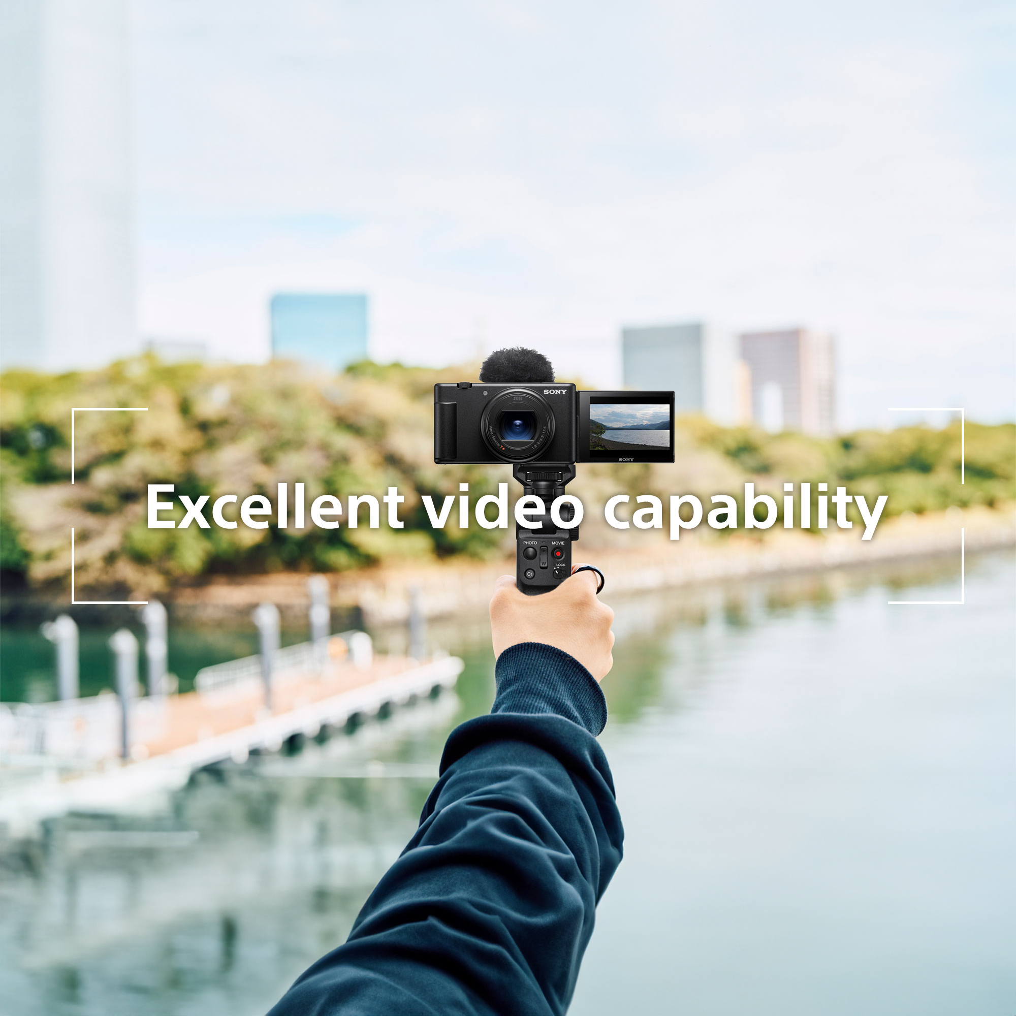 Eine Hand hält eine kompakte Videokamera mit Stativ vor einer Uferlandschaft mit Fluss und Stadt.