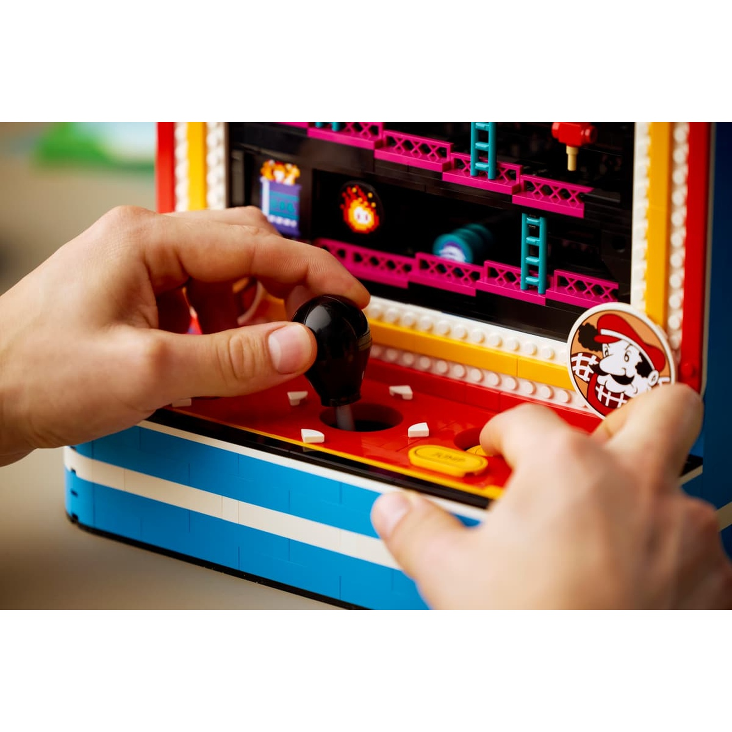 Mani che giocano con un colorato cabinato arcade Lego con joystick e pulsanti.