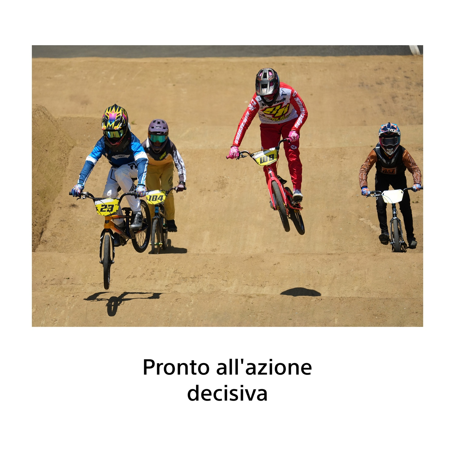 Quattro ciclisti BMX corrono su una pista sterrata, uno in aria con abiti rossi.