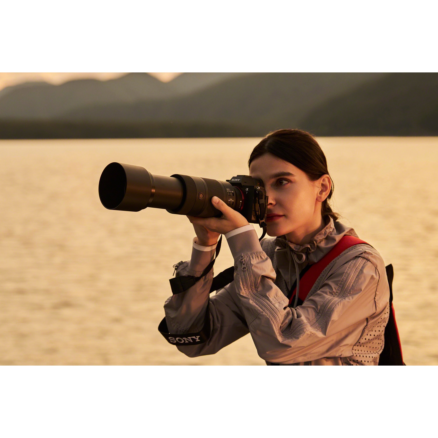 Una donna fotografa con una fotocamera Sony e un grande teleobiettivo davanti a un paesaggio fluviale e montano al tramonto.