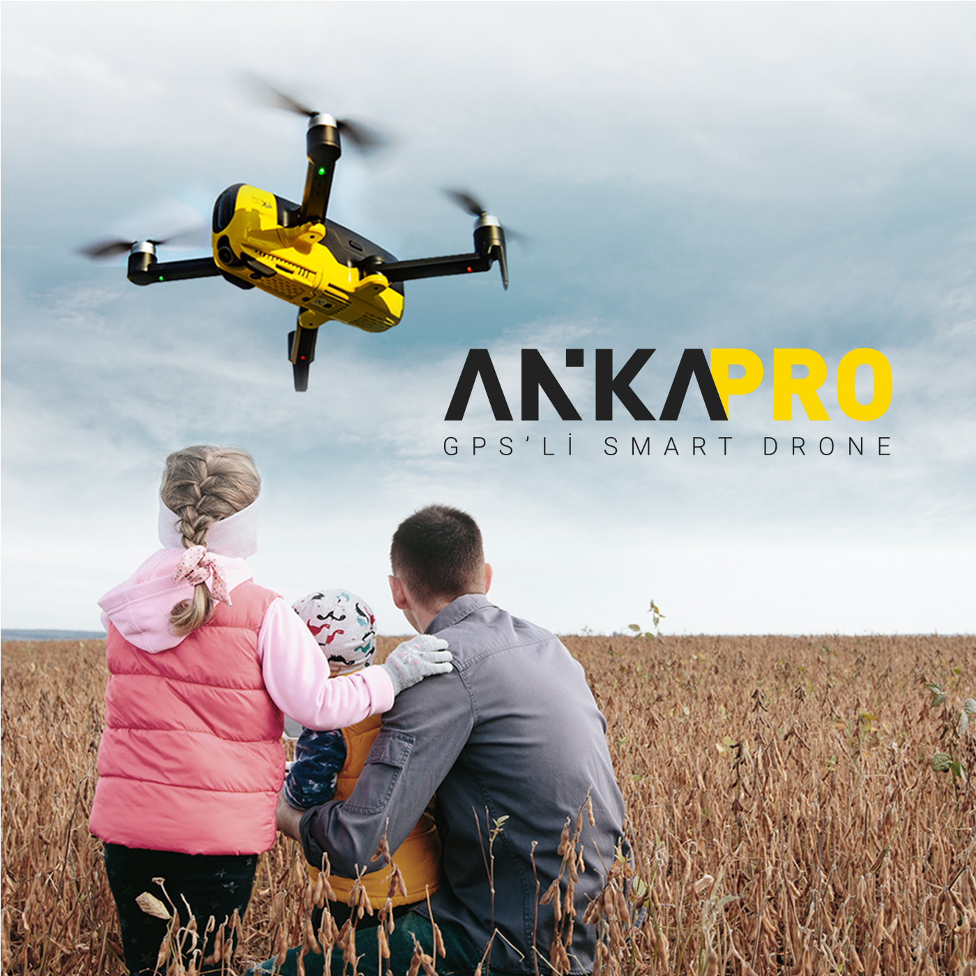 Bir aile, ANKAPRO yazılı hasat edilmiş bir tarlanın üzerinde sarı bir dron izliyor.