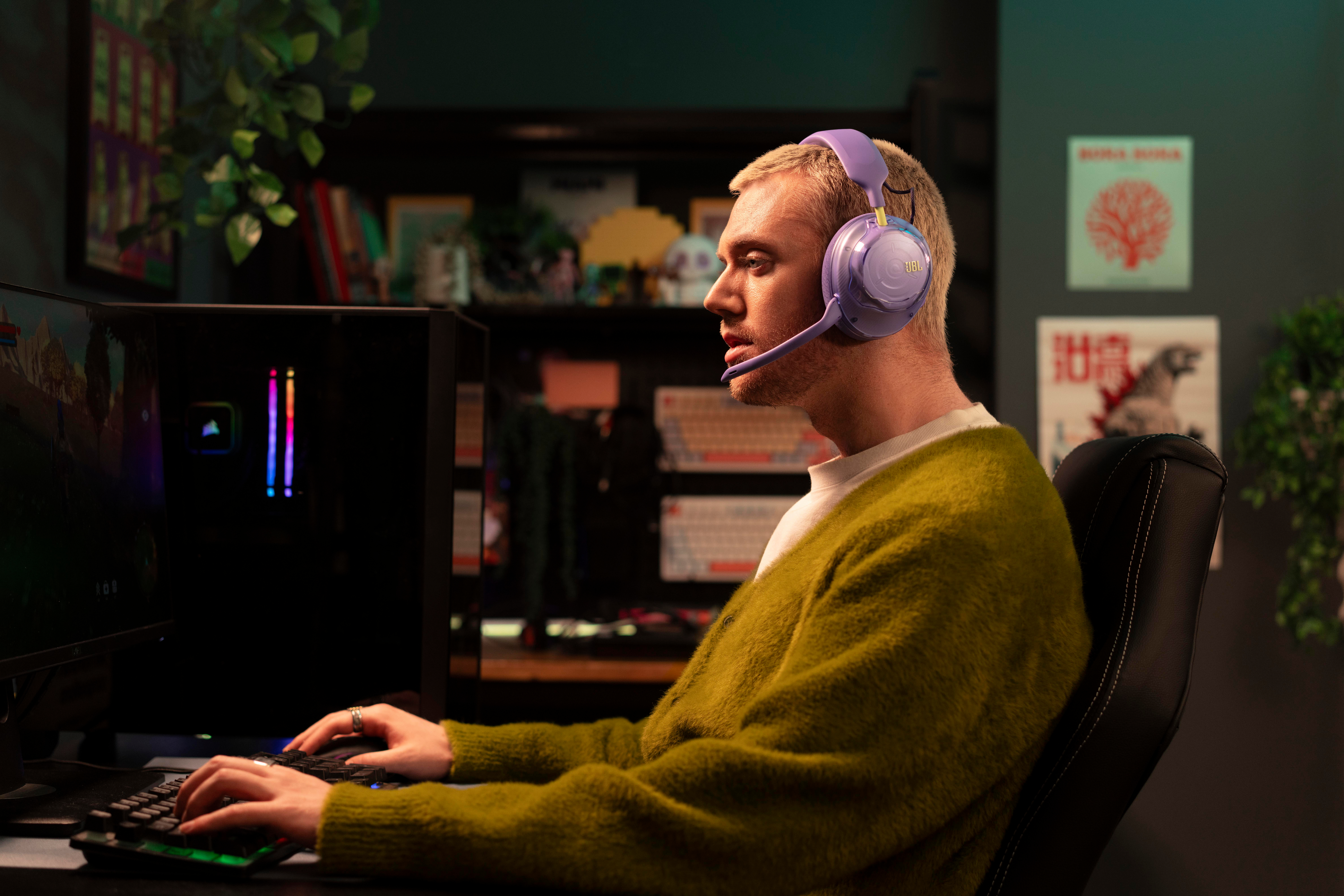 Ein Mann im grünen Pullover und mit violetten Kopfhörern sitzt am Computer und tippt.