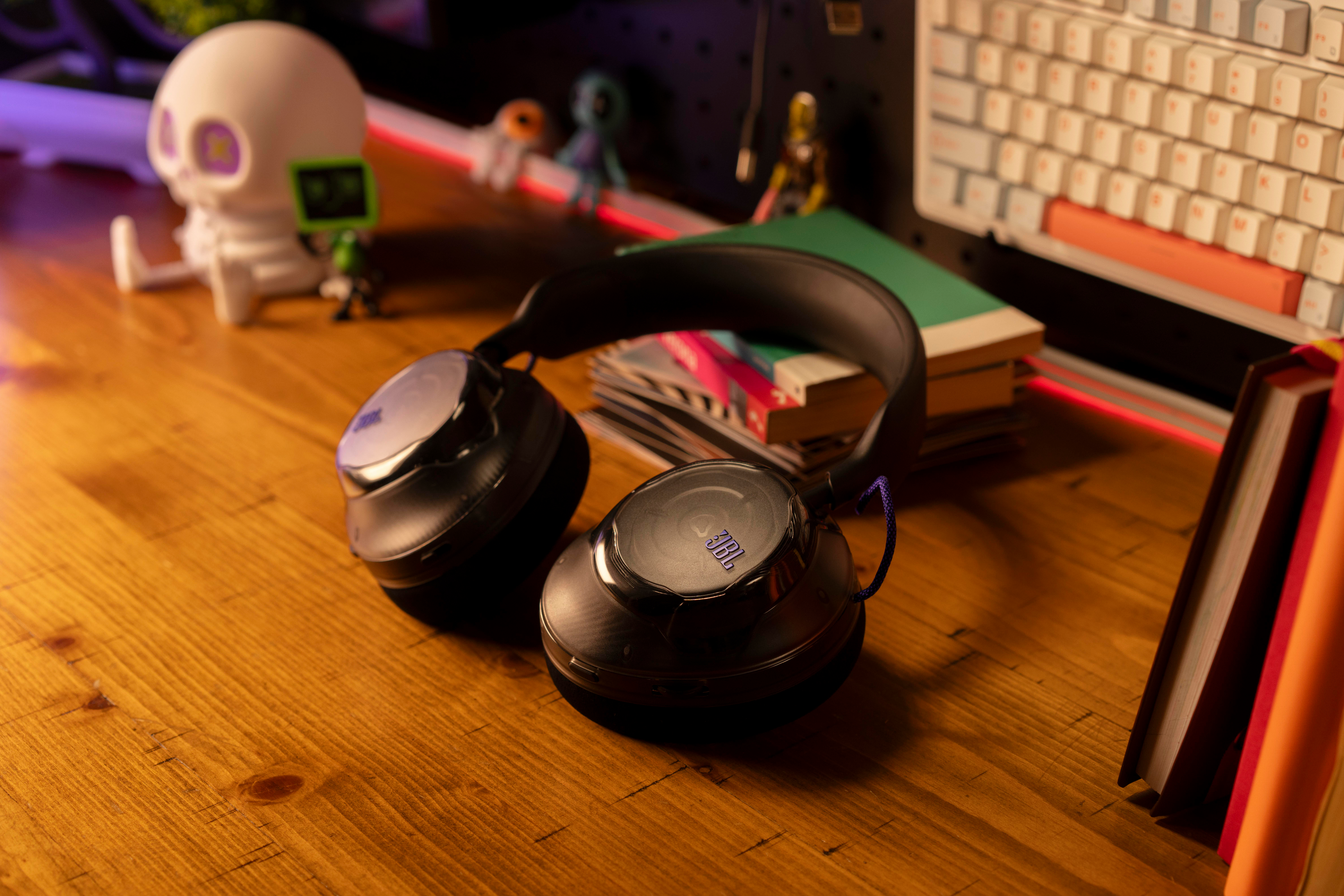 Schwarze JBL Over-Ear-Kopfhörer auf einem Holztisch mit Büchern, einer Tastatur und einer Totenkopf-Figur im Hintergrund.