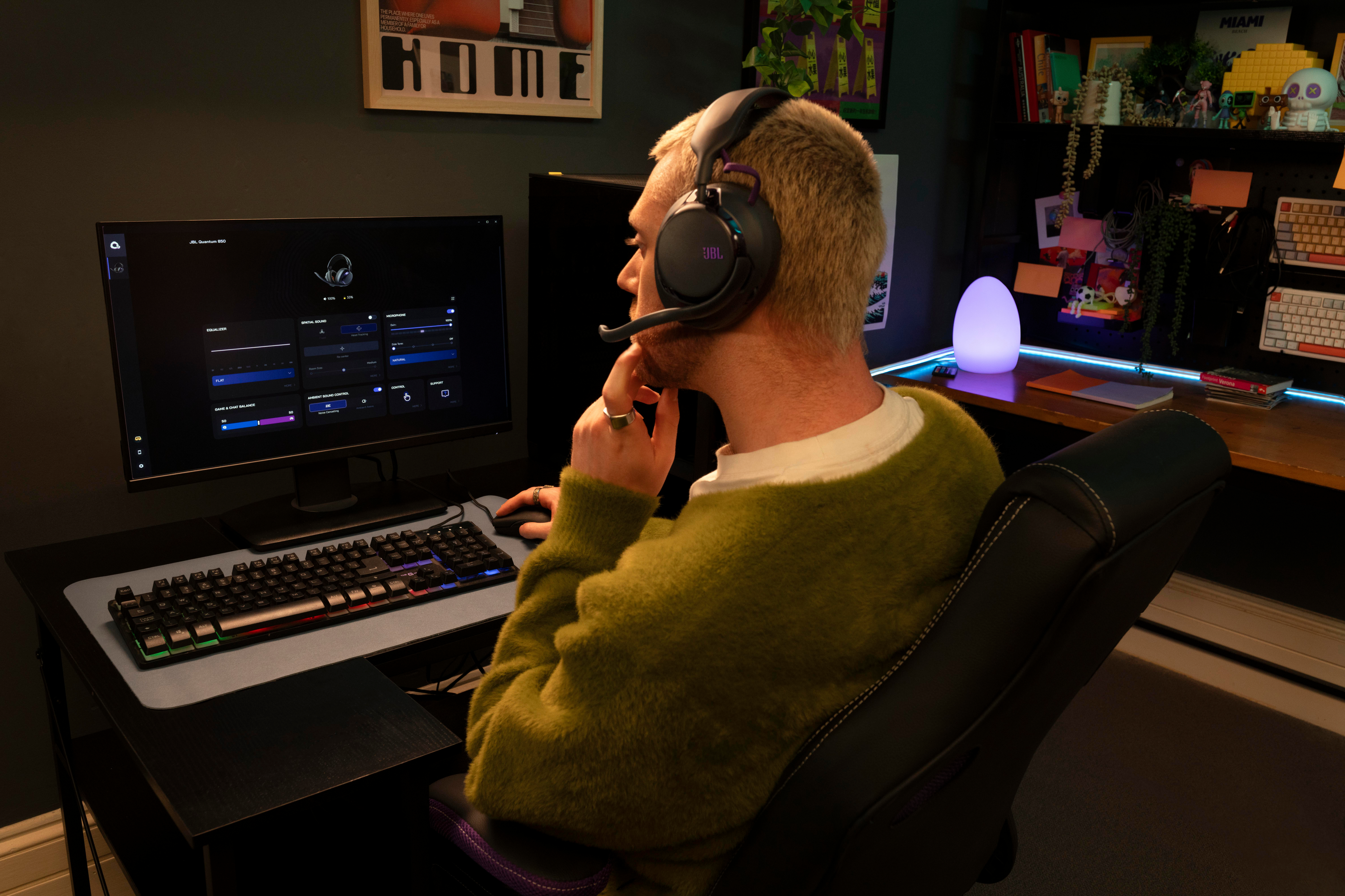 Ein Mann im grünen Pullover mit Headset sitzt am Schreibtisch und schaut auf einen Monitor.