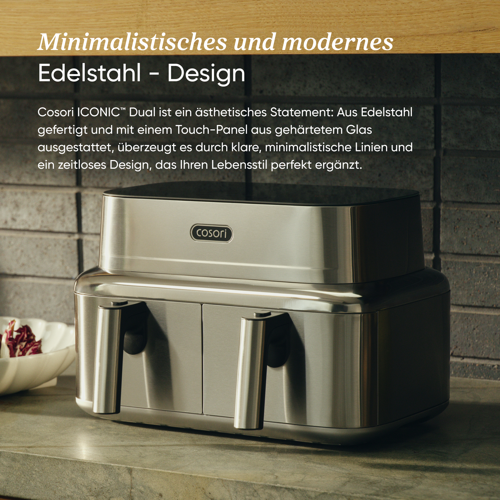 Moderne Edelstahl-Heißluftfritteuse von Cosori auf einer Arbeitsplatte vor grauer Ziegelwand.