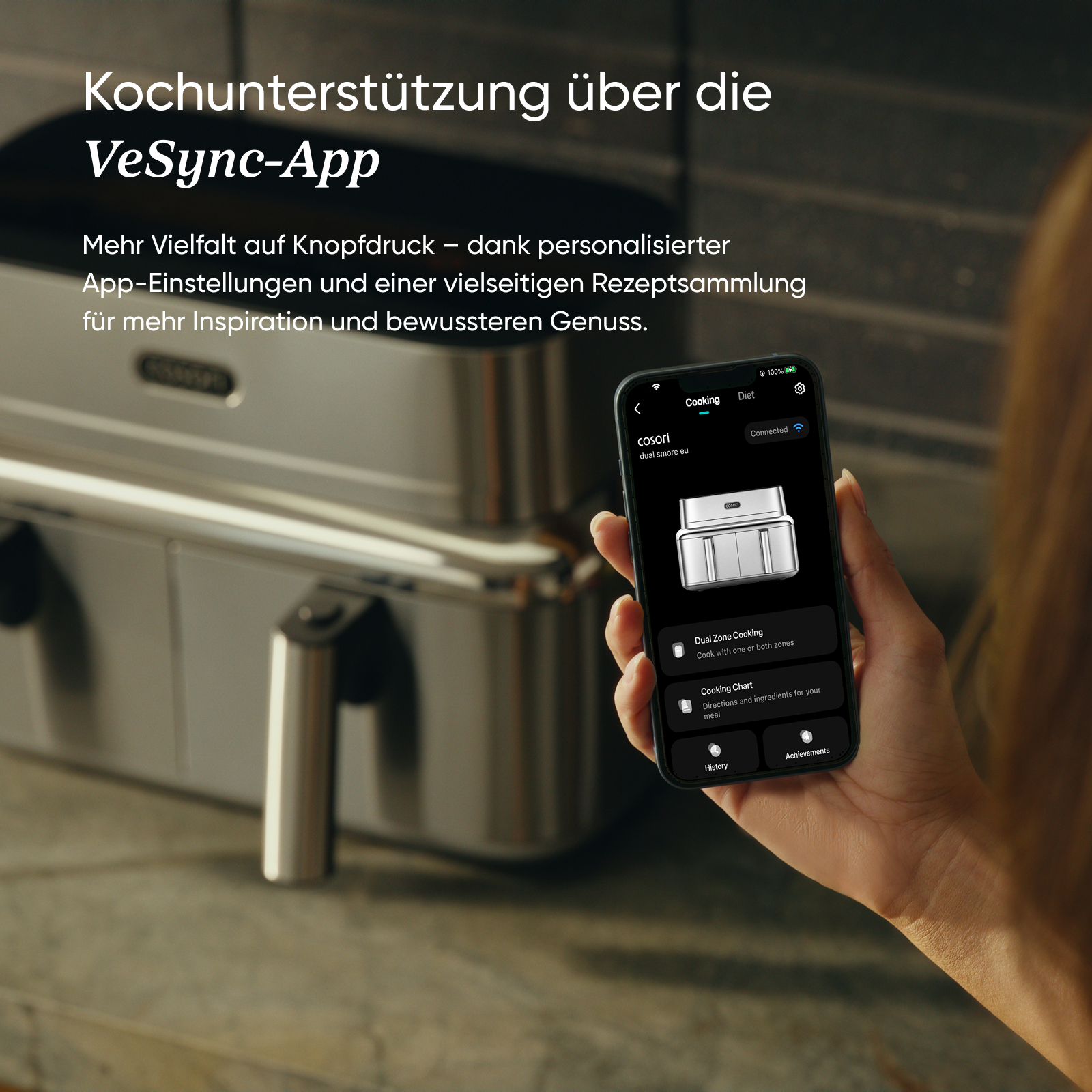 Eine Person hält ein Smartphone mit der VeSync-App, im unscharfen Hintergrund eine Heißluftfritteuse.