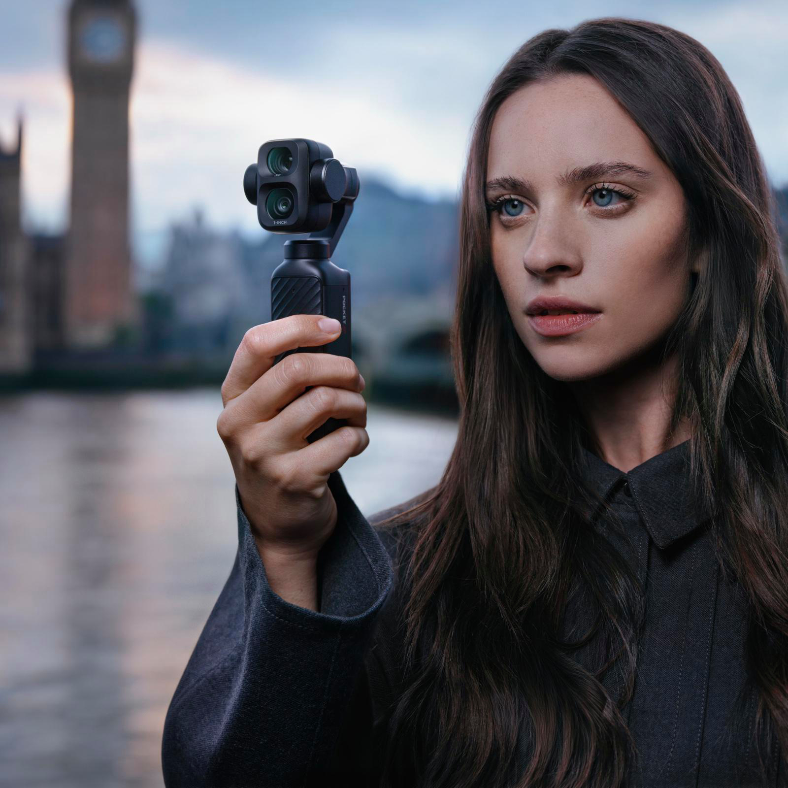 Eine Frau hält eine kleine schwarze Gimbal-Kamera in der Hand, im Hintergrund eine Flusslandschaft mit Turm.