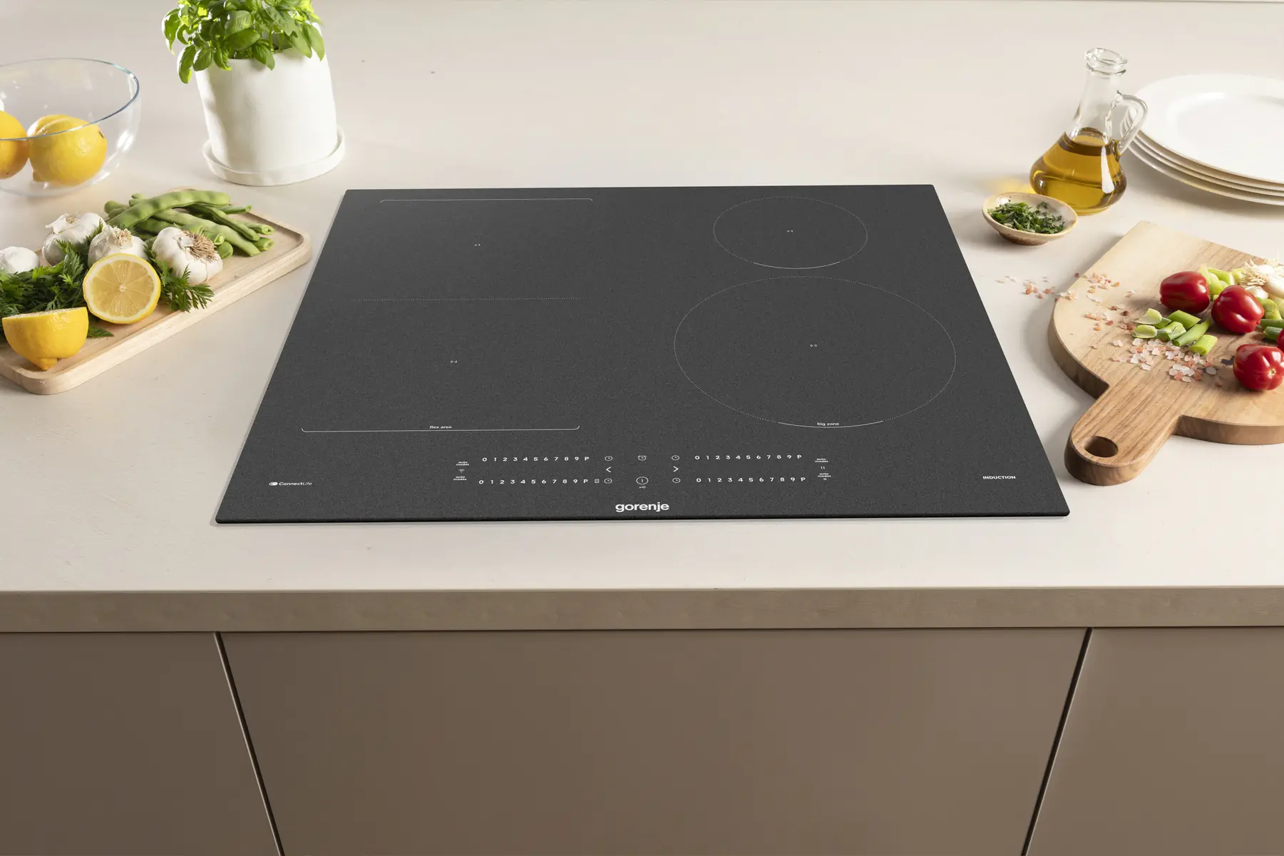 Egy fekete Gorenje indukciós főzőlap konyhapulton, zöldségvágó deszkákkal és olajjal körülvéve.