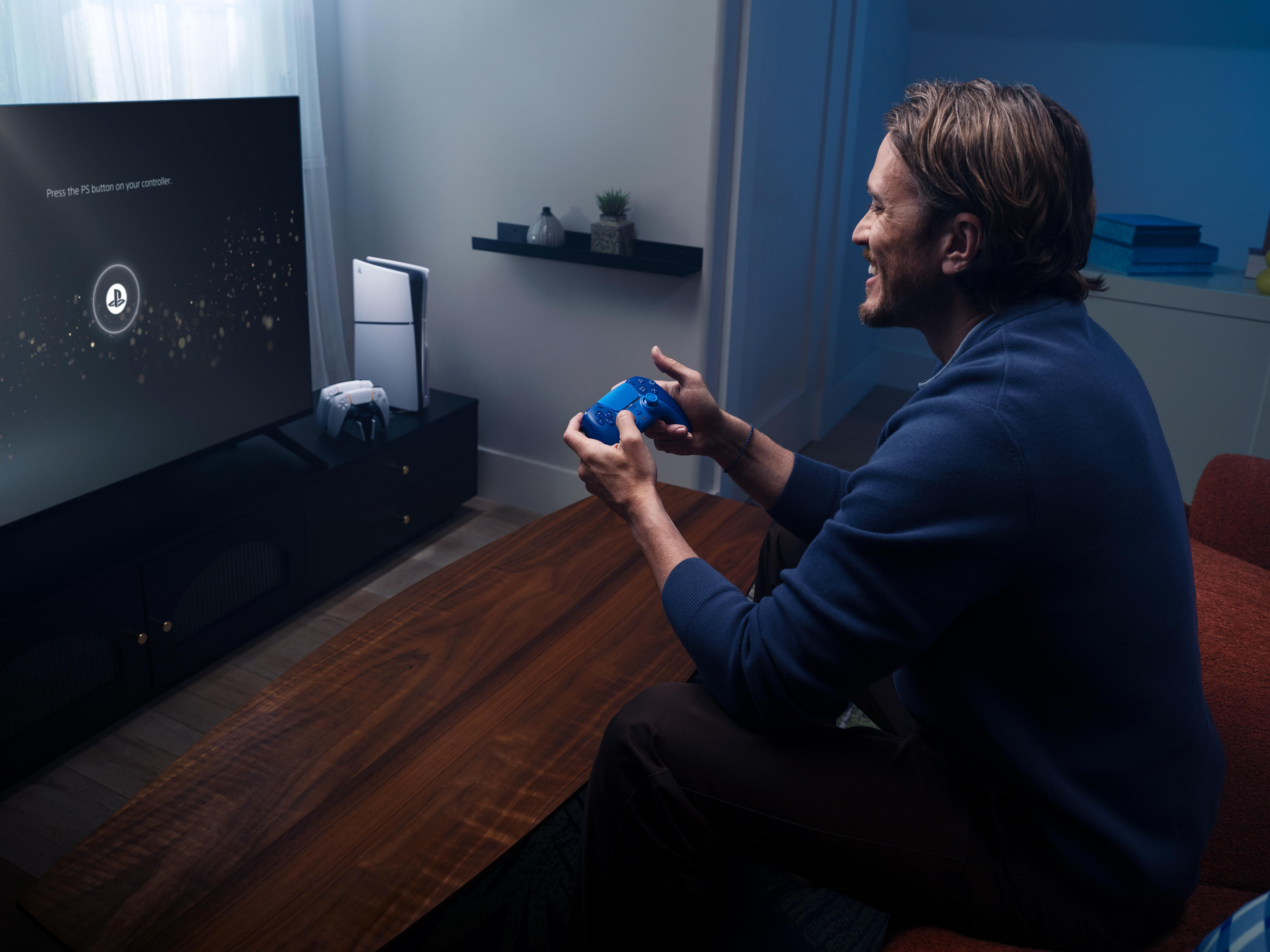 Ein Mann lächelt, während er in einem Wohnzimmer mit einem blauen Controller ein Videospiel auf dem Fernseher spielt.