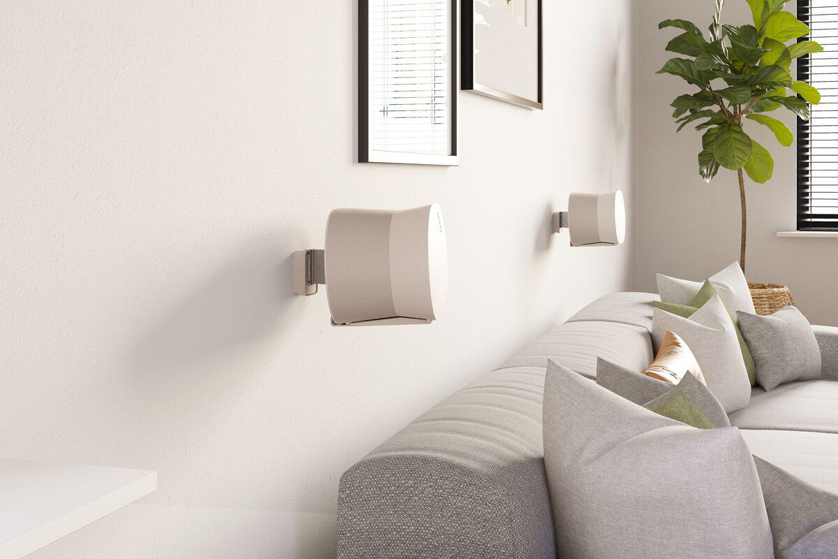 Wohnzimmer mit grauer Couch, Kissen, Wandlautsprechern, gerahmten Bildern und grüner Topfpflanze.